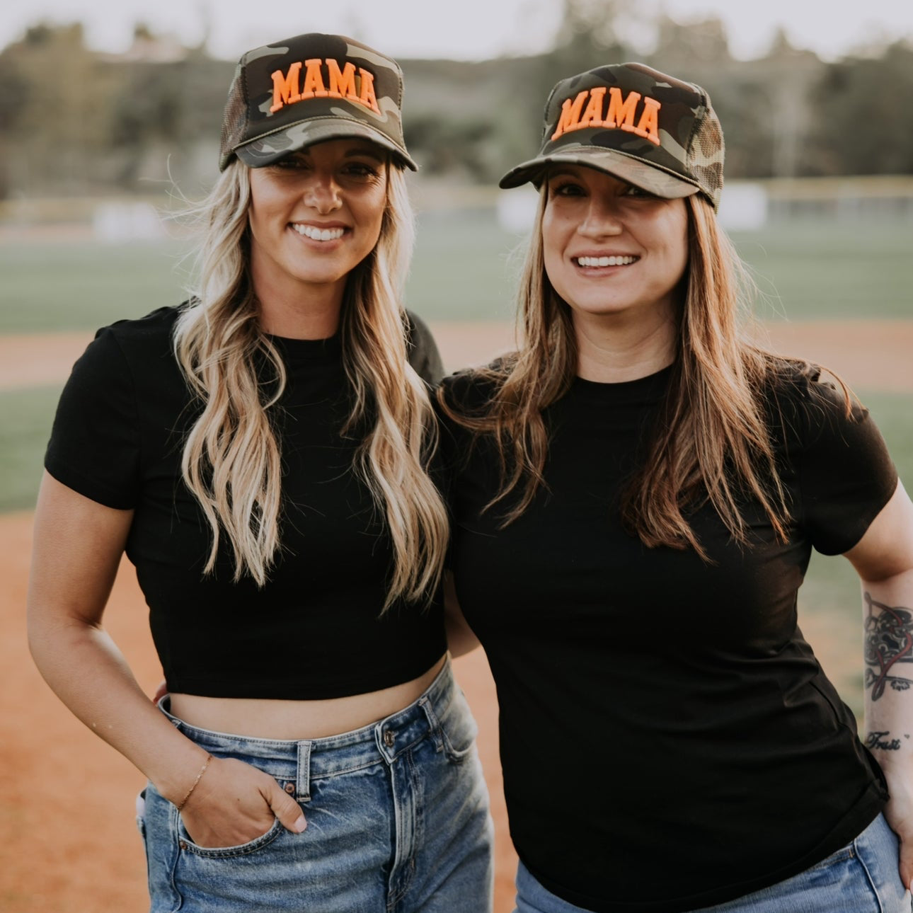 Two women stand on a baseball field, smiling wide. They're wearing matching Foam Camo Neon Mama Trucker hats from Mama X™ Brand, paired with black t-shirts and blue jeans—a perfect photo op against the trees and baseball backdrop, ideal for Mother’s Day gifts.