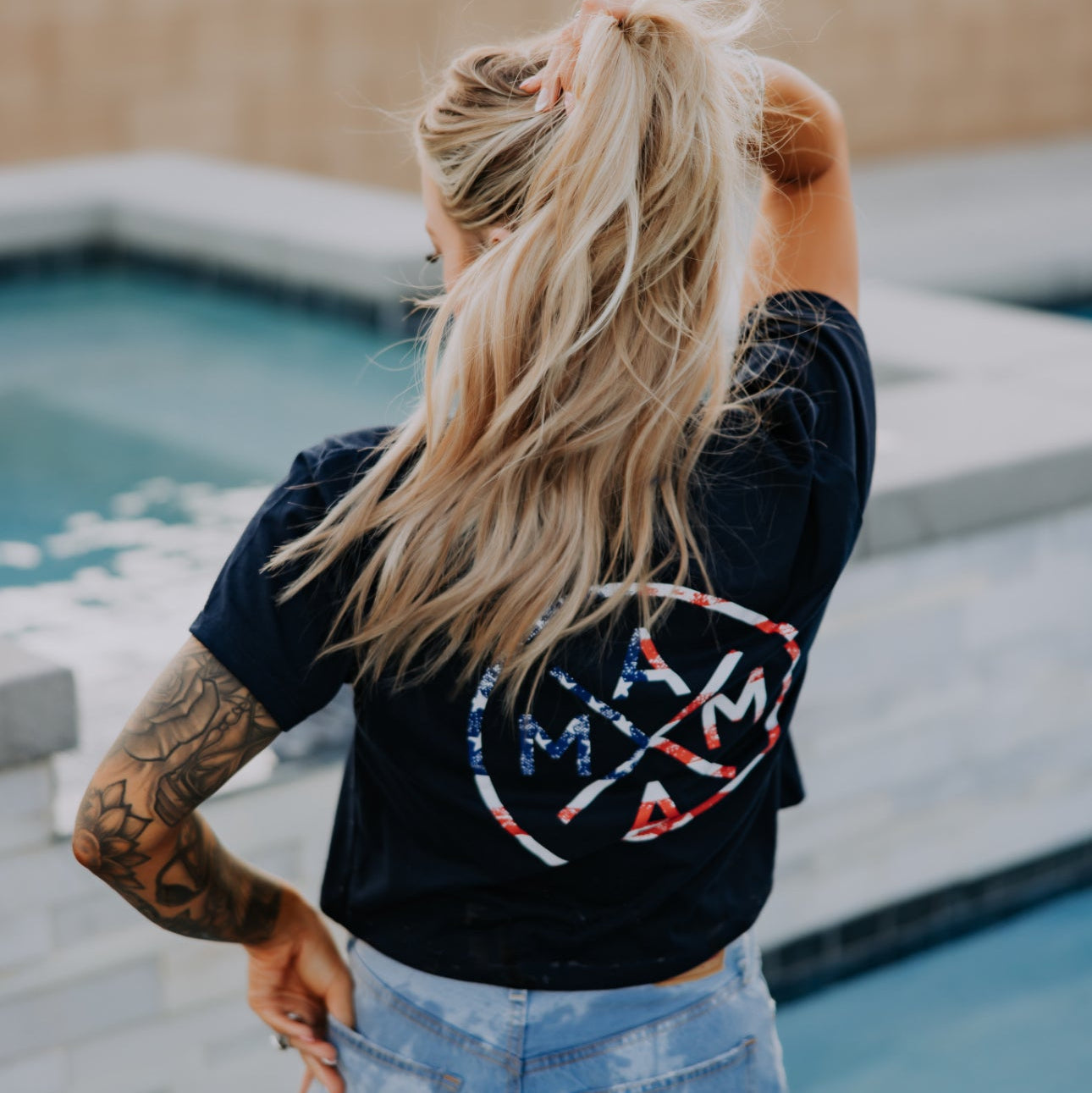 A woman with long blonde hair in a ponytail stands by a pool, back to the camera, wearing the Mama X™ Brand’s Mama X Flag Crop Top and denim shorts—showing off arm tattoos for a perfect summer look.