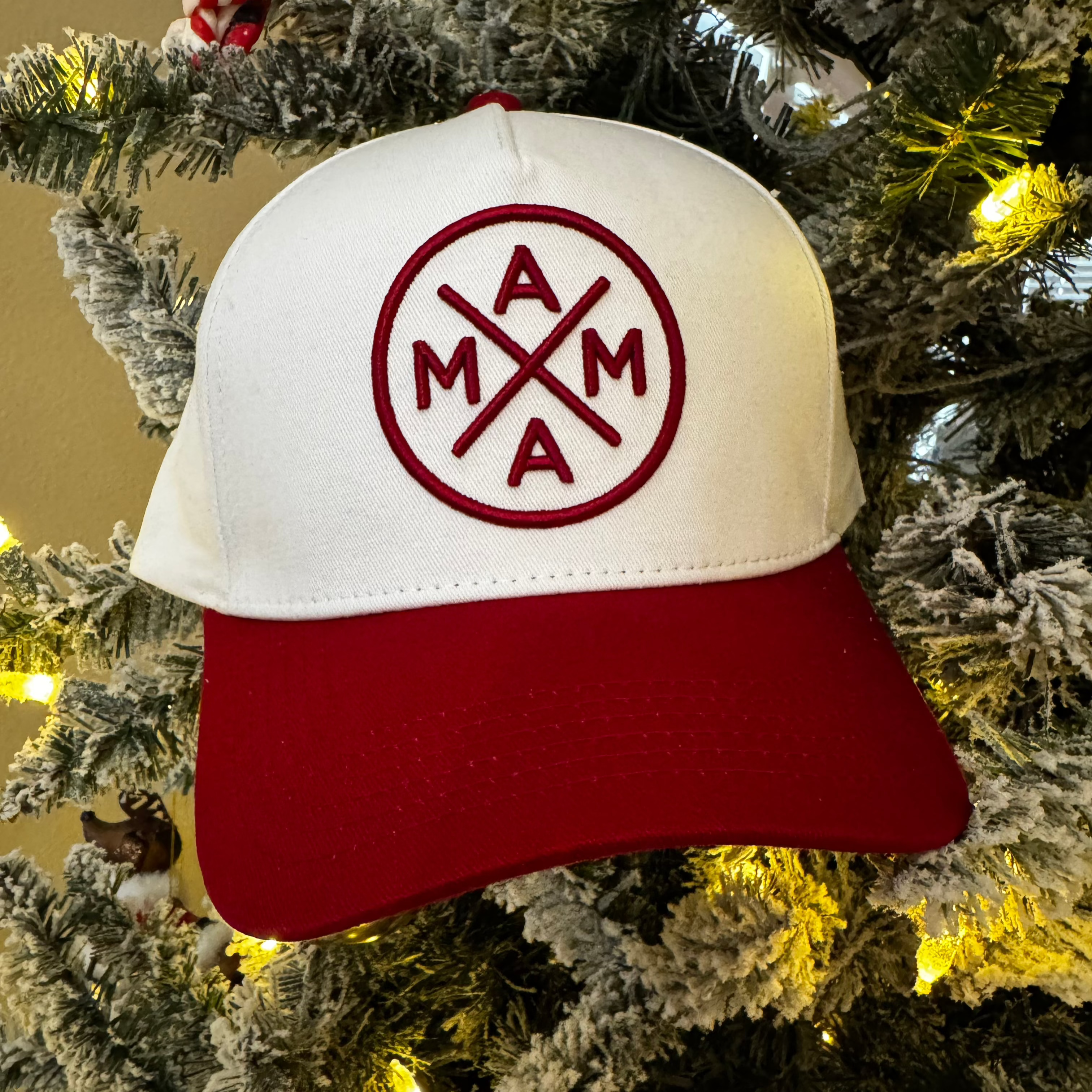 The Red Mama X™ Baseball Cap by Mama X™ Brand, featuring a white and red design with the letters "MAMA" arranged in a circular cross pattern on the front, is displayed atop a Christmas tree dusted with snow and decorated with yellow lights and small ornaments.