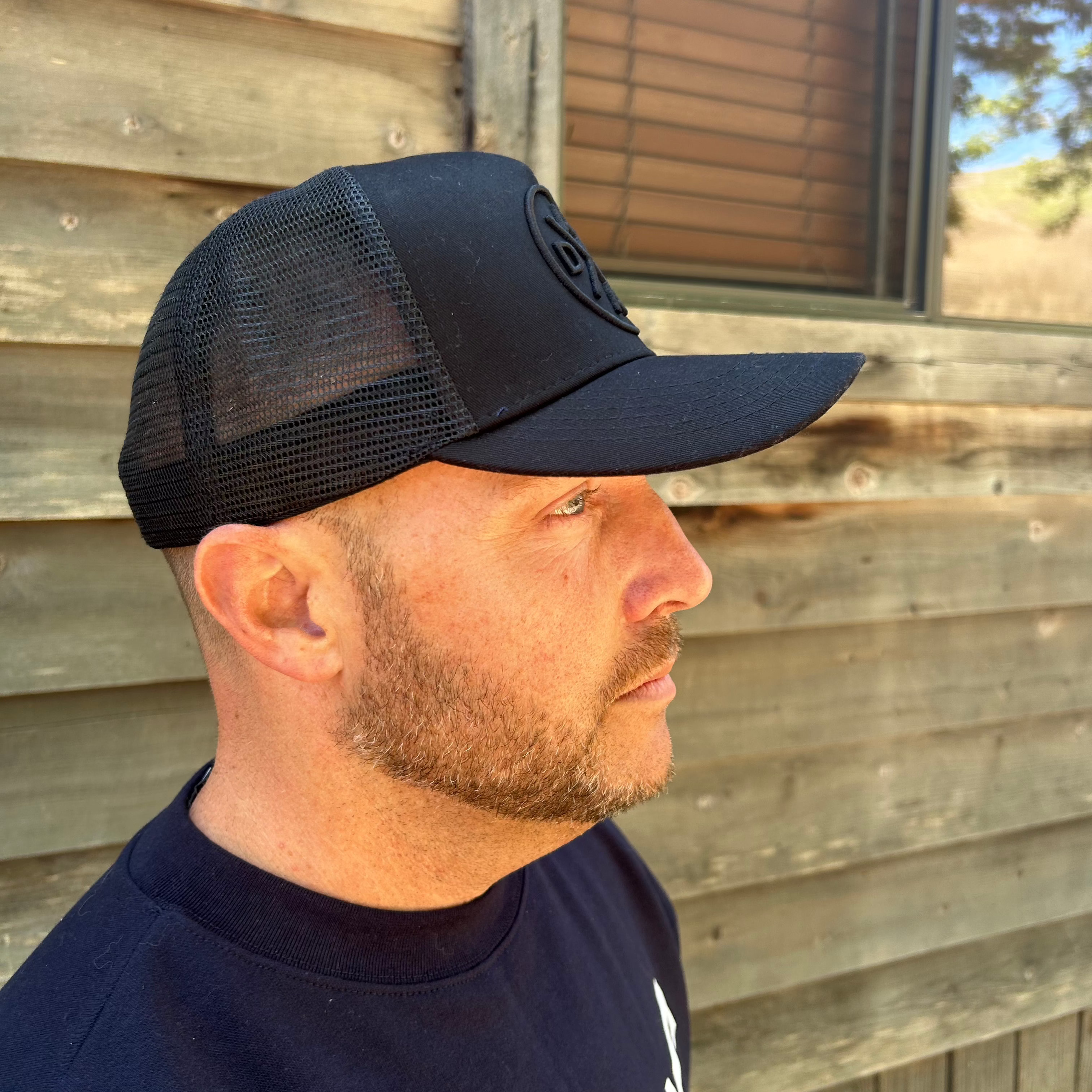 A man with a short beard is wearing the Dada X™ Blackout Premium Trucker Hat with a curved brim from Mama X™ Brand, featuring a 3D embroidered logo. He is facing slightly to the left, with a textured wooden wall and small window in the background. The scene appears to be outdoors in daylight.