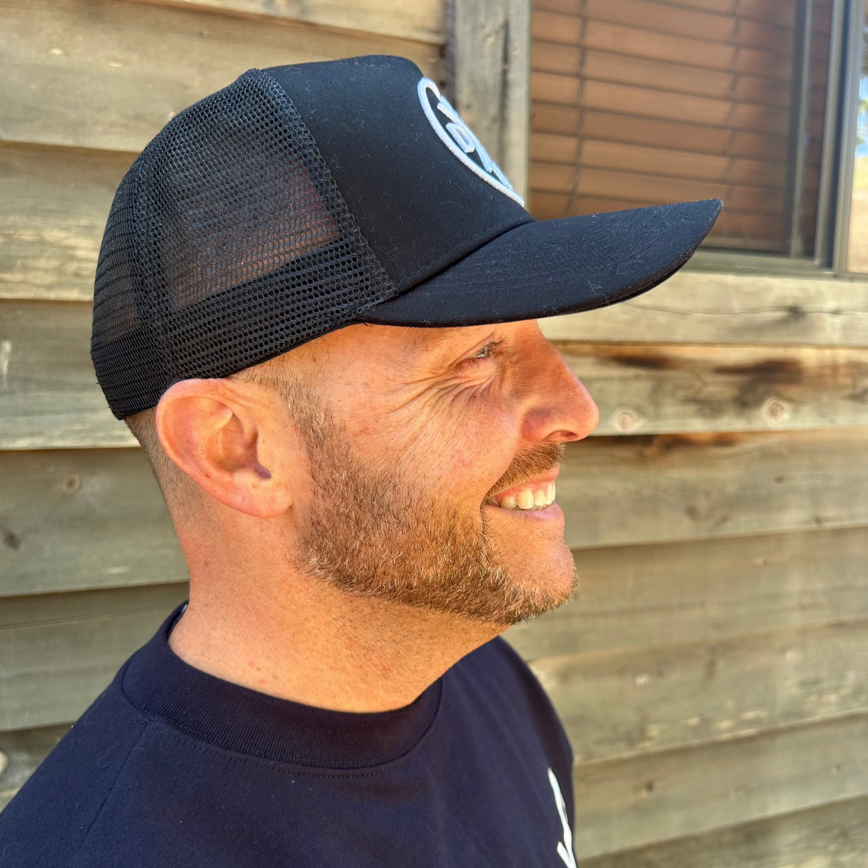 A bearded man smiles while looking to the right, wearing a Dada X™ Premium Trucker Hat Curved Brim from Mama X™ Brand and a dark crew-neck shirt. The background features a wooden wall with a window.