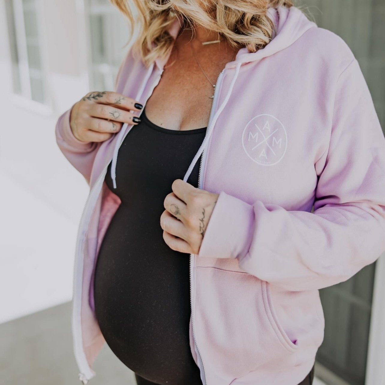 A pregnant woman showcases her baby bump while wearing a Mama X™ Zip Up Sweatshirt in lavender and an open pink hoodie that reads "MAMA." She has shoulder-length wavy blonde hair and stands indoors near a window, holding the sides of her premium cotton blend hoodie with her hands.