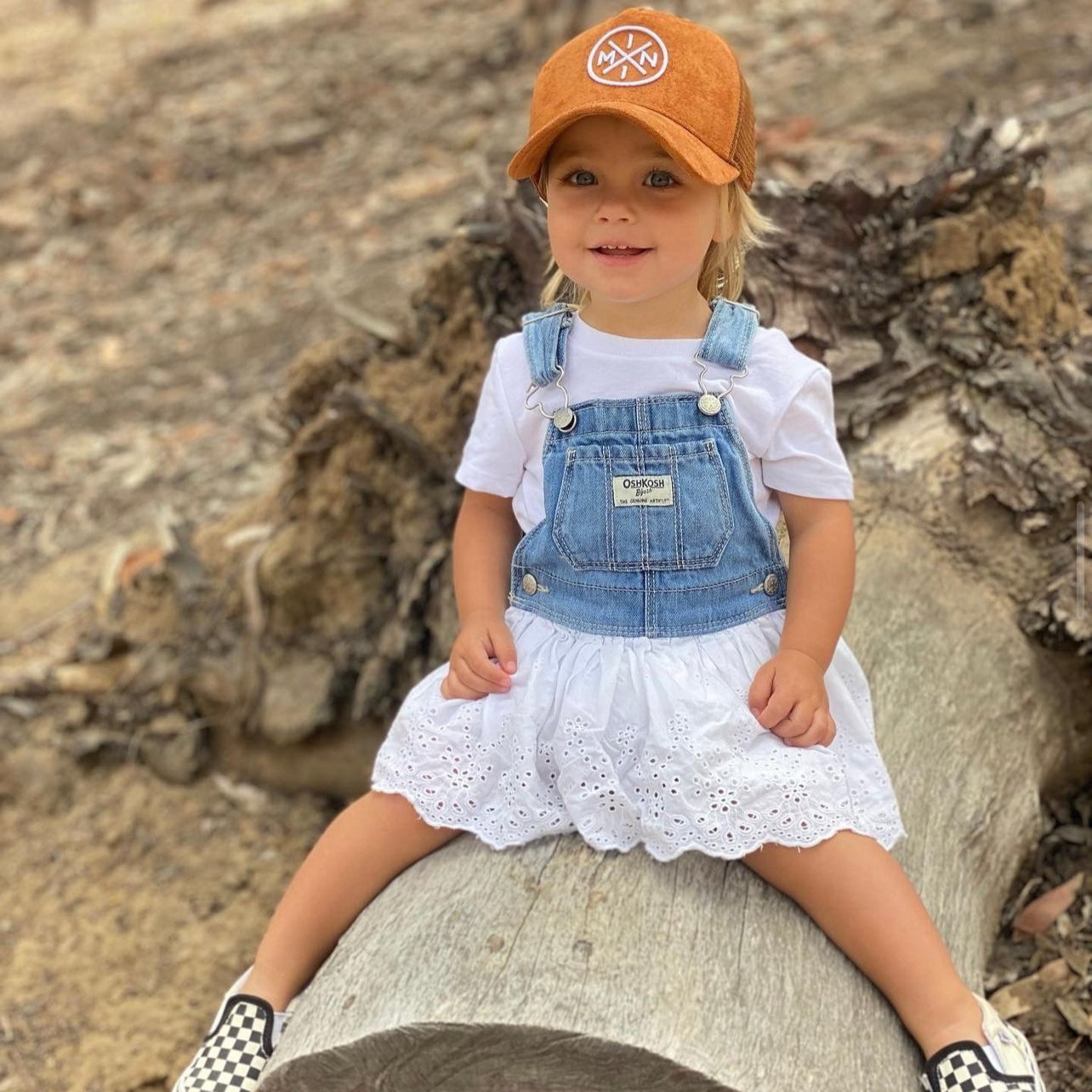 A young child sits on a large log outdoors, wearing the cutest hat—a Mini X Brown Suede Trucker Hat from Mama X™ Brand—paired with a white t-shirt, denim overalls with a white lace skirt underneath, and black-and-white checkered slip-on shoes. The background reveals a natural setting with dried foliage and wood, perfect for a fall outfit.
