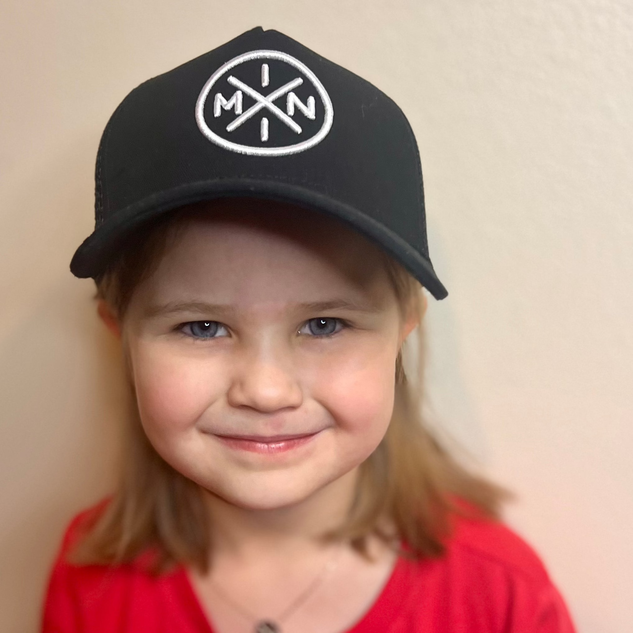 A young child with light brown hair smiles while wearing the Mini X Premium Trucker Hat from Mama X™ Brand. The black cap is adorned with a circular white embroidered logo featuring the letters "M," "I," and "N." The child, dressed in a red shirt, stands against a plain beige background.