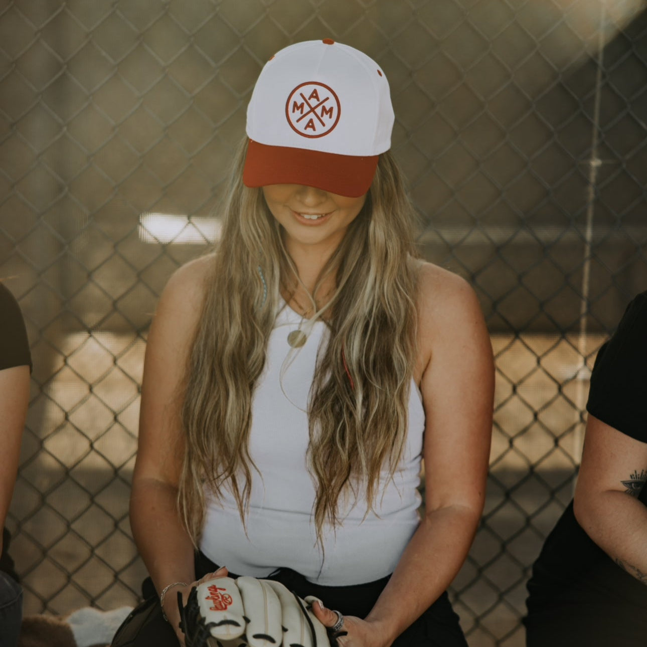 Wearing a Red Mama X™ Baseball Cap from the Mama X™ Brand, a long-haired individual in a white tank top sits on a bench, holding a baseball glove. They gaze downward, lost in thought, surrounded by nostalgia with a chain-link fence as their backdrop.