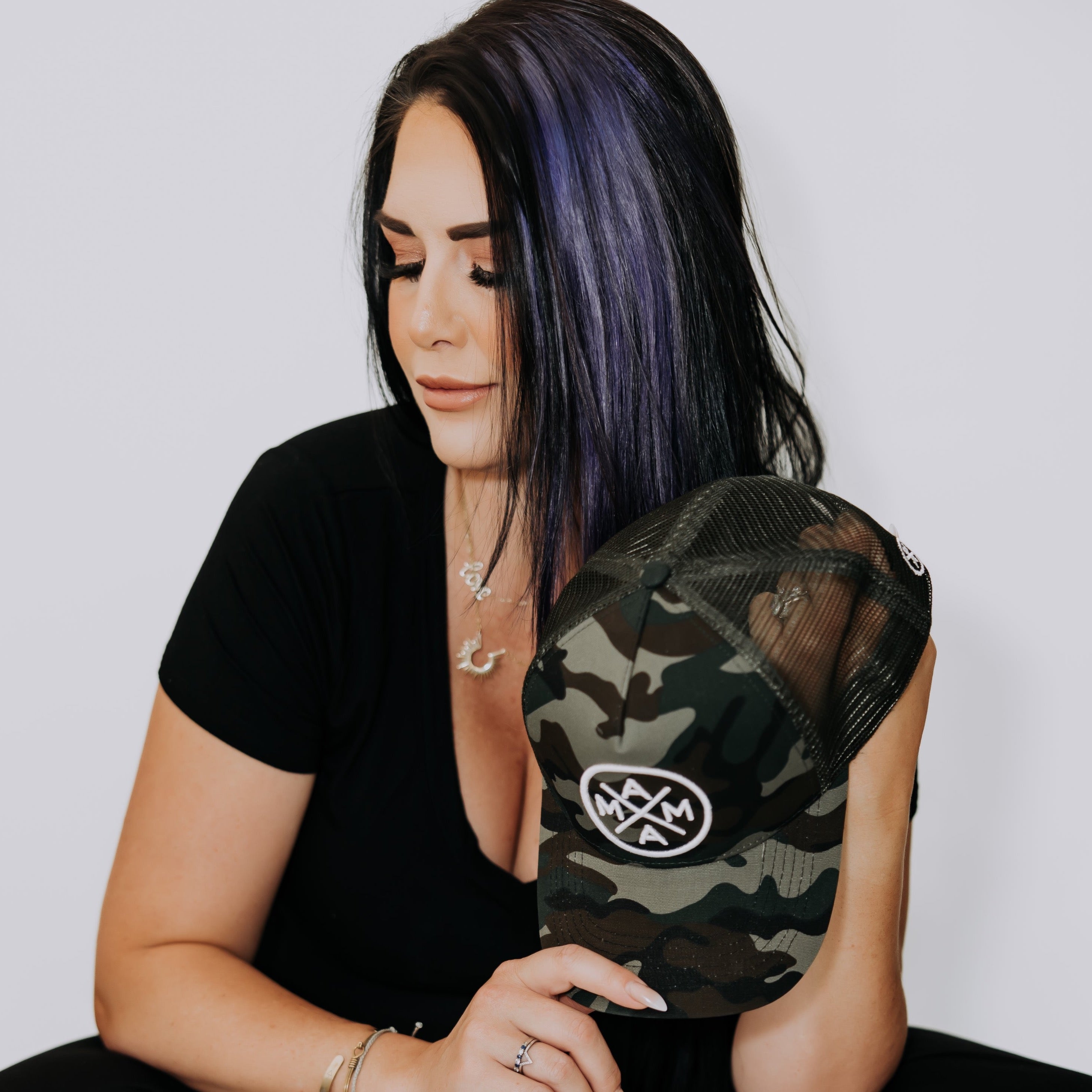 A person with long, dark hair subtly highlighted in purple gazes downward with a serene expression. They hold the Mama X™ Premium Canvas Trucker Hat in Camo from the Mama X™ Brand while wearing a black V-neck top and several bracelets on their wrist. The background is plain white.