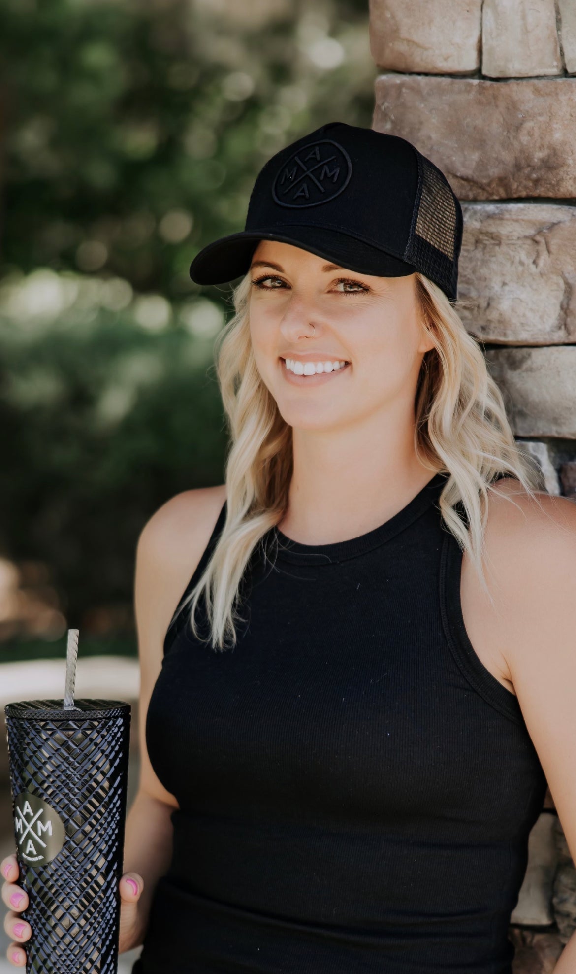 A woman with long blonde hair is smiling while leaning against a stone wall. She is wearing a black tank top and a Mama X™ Brand Blackout Premium Trucker Hat. She holds a black textured cup with a straw. The background is green and blurred, suggesting an outdoor setting.