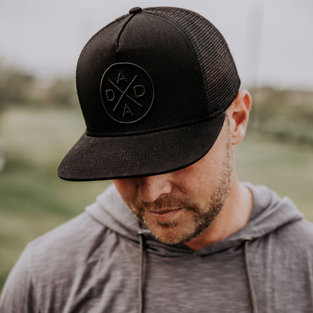 A person with a beard is looking down while wearing the Dada X™ Blackout Premium Trucker Hat from Mama X™ Brand and a grey hoodie. The background is blurred, giving the impression of an outdoor setting. The hat features a circular stitched 3D logo with the word "DADA" arranged in a cross pattern.