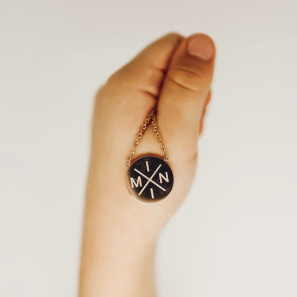 A hand holding the Mama X™ Brand Mini X Necklace in rose gold, featuring a round pendant with a black background. The letters "M," "I," "N," and "I" are arranged in a cross pattern, with a small "X" at the intersection of the lines. This stylish yet durable accessory is perfect for children&