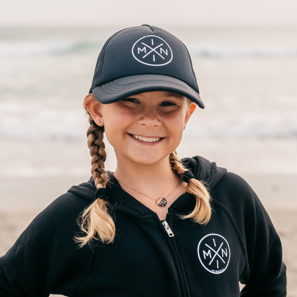 A cheerful young girl with braided blonde hair smiles at the camera, her black cap and black hoodie with matching intersecting letter logos lending a stylish touch. She proudly sports the Mini X Necklace from Mama X™ Brand, its rose gold gleam standing out against the blurred ocean in the background.
