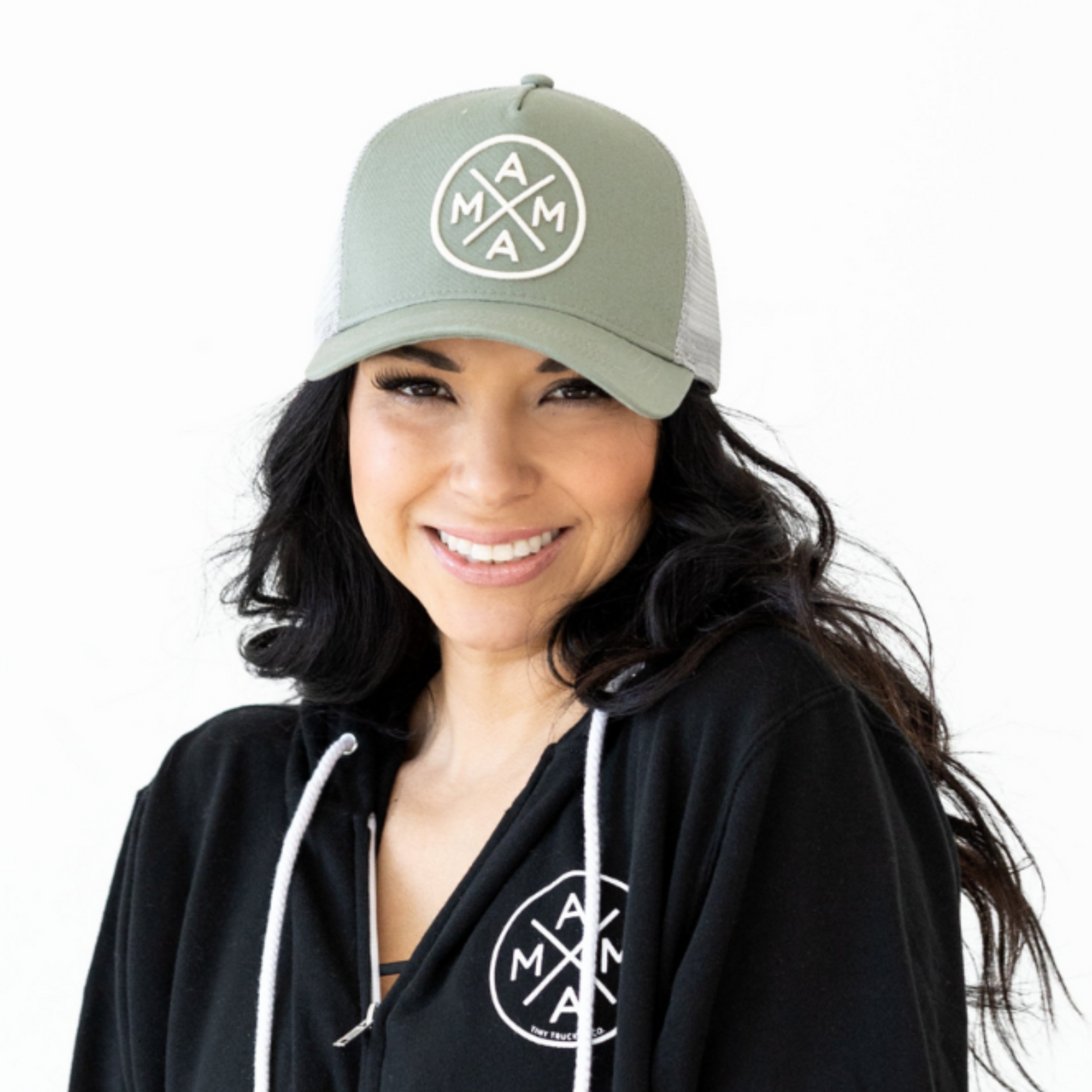 A smiling woman wearing the Mama X™ Premium Canvas Trucker Hat in sage and a black hoodie adorned with the circular MAMA X™ logo. The 5-panel cap features a mesh back, and her long, dark hair flows over her shoulders as she stands against a plain white background.