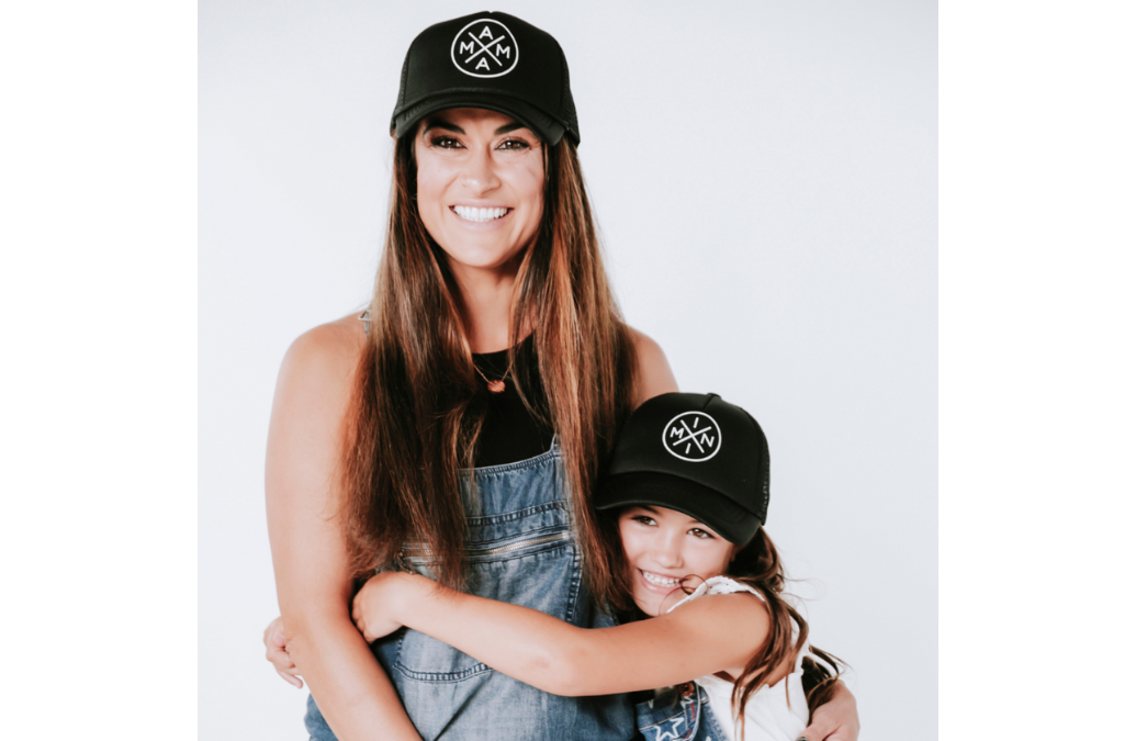 A woman and a young girl, both smiling at the camera, are wearing the best-seller **Mama X™ Foam Trucker Hat** by **Mama X™ Brand**. The woman, with long brown hair, is dressed in denim overalls while the girl, who also has brown hair and is wearing a sleeveless dress, gives her a hug from the front. The background is plain white.