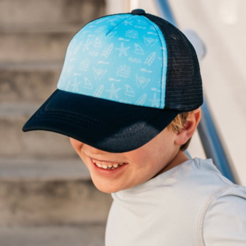 A smiling child wearing a Mama X™ Brand Shaka Repeater Trucker Hat, which features blue and black 5-panel construction with beach-themed illustrations on the front, stands near a staircase. The white shirt contrasts with the hat's brim that partially shades the child's face. The plastic adjustable snap at the back ensures a perfect fit.