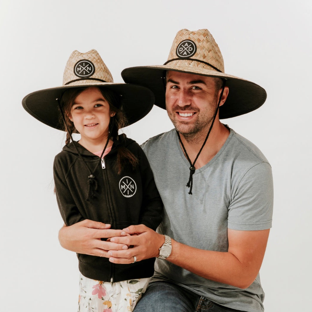 A smiling man and a young girl pose together, both wearing large, stylish Dada X™ Lifeguard Hats with wide brims and the Mama X™ Brand's black logo on the front. The man is dressed in a grey t-shirt and jeans, while the girl sports a black hoodie and patterned skirt. They stand against a plain, white background.