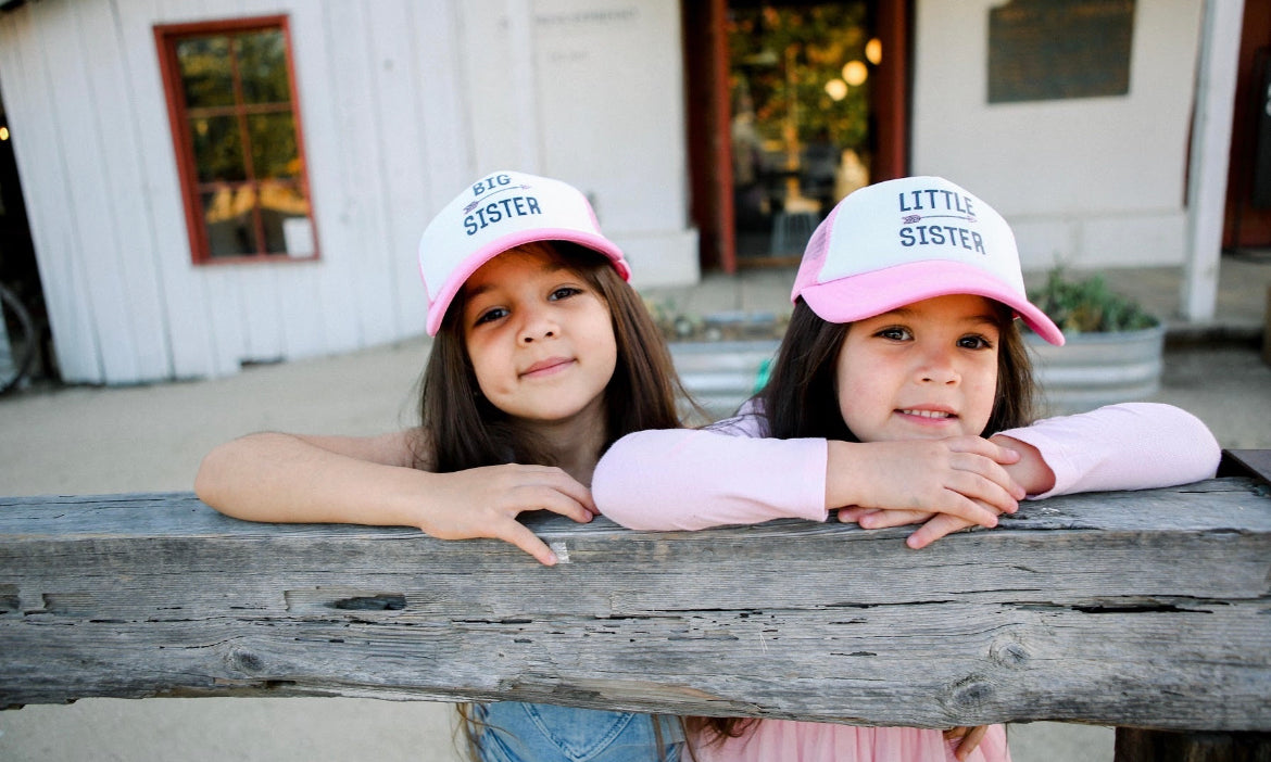 Two young girls lean on a wooden fence. Both are wearing caps; one is labeled "BIG SISTER," while the other is a light pink trucker hat from Mama X™ Brand, labeled "Little Sister Trucker Hat." They smile at the camera with a rustic wooden building and potted plants in the background.