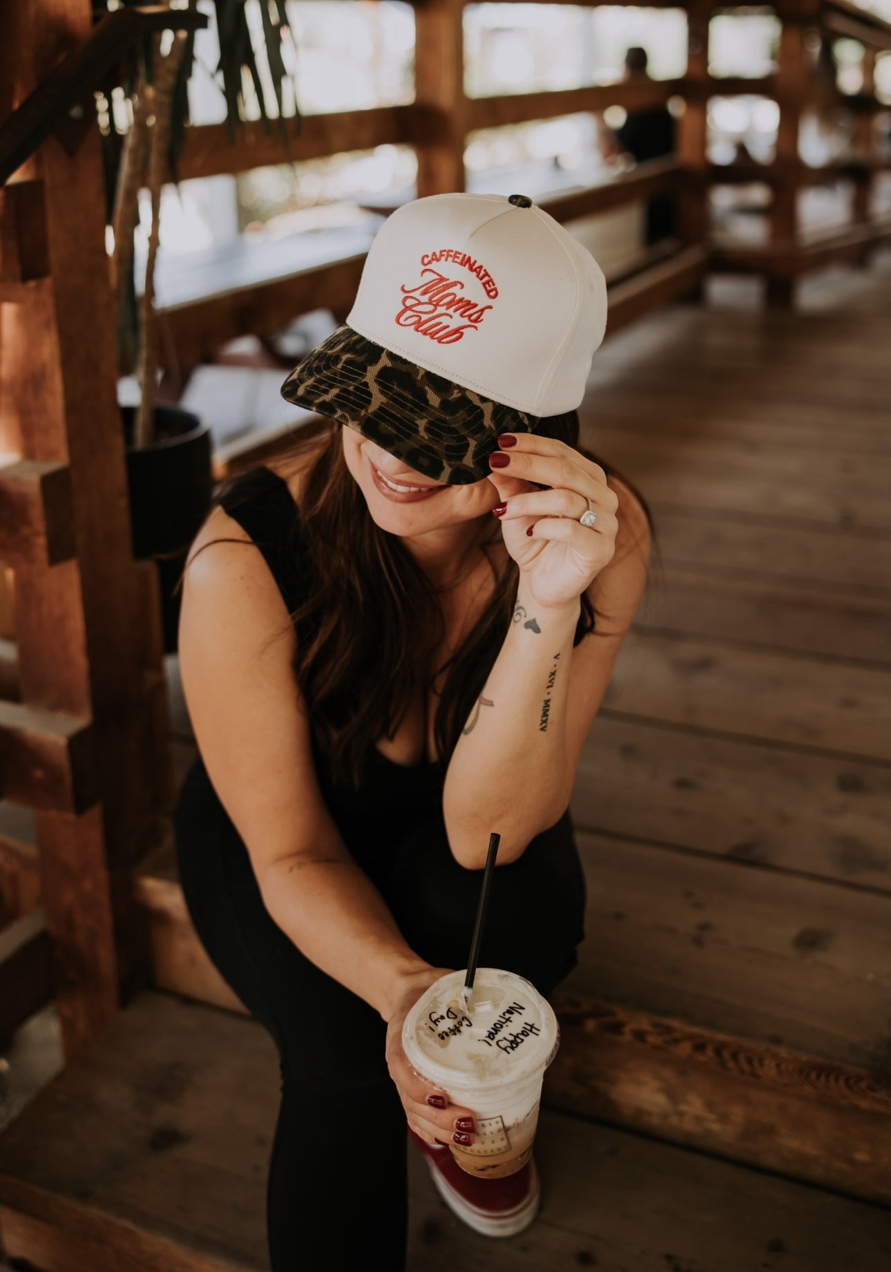 A woman walks up wooden stairs, wearing a green shirt with bold red text on the back that reads, “BRING ME AN ICED COFFEE.” Her hair is tied back with a scrunchie.