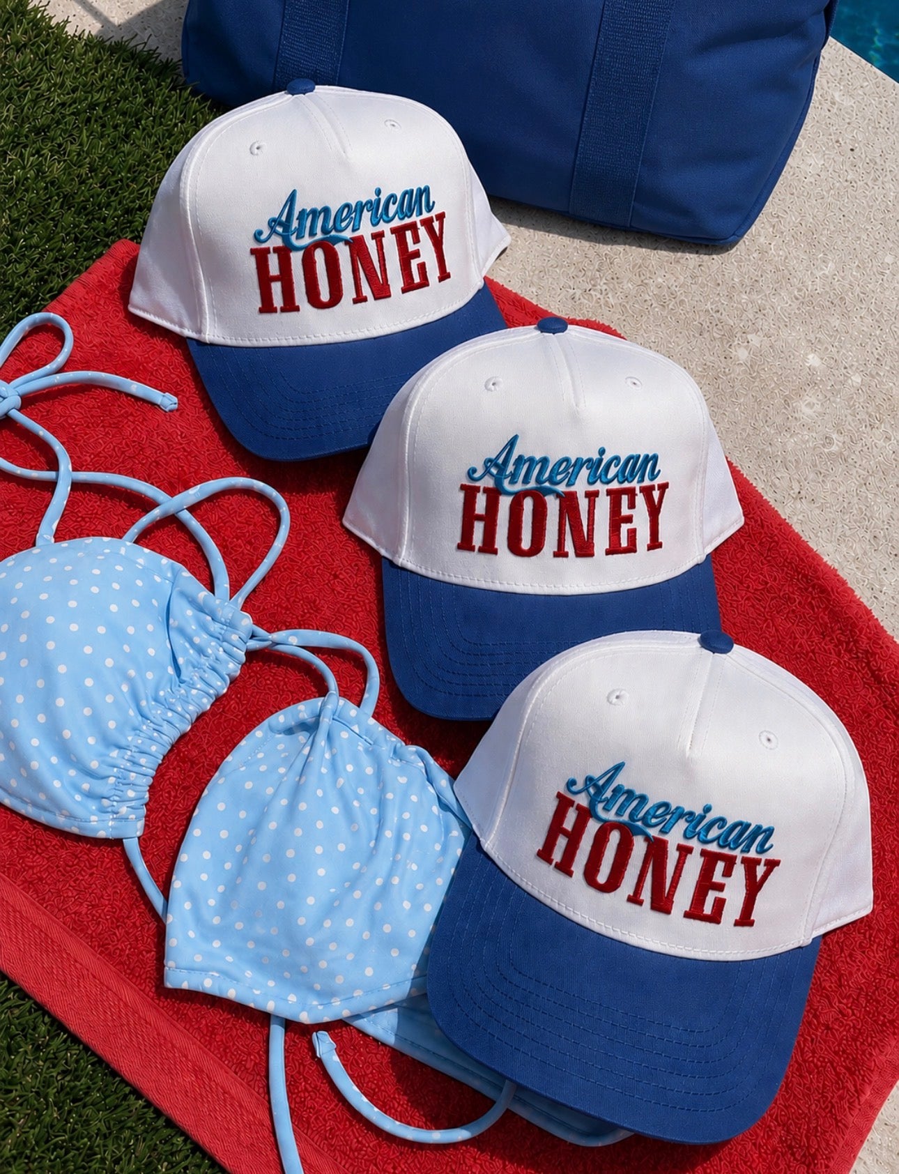 Three white and blue baseball caps with "American HONEY" written on them are placed on a red towel next to a light blue polka dot bikini and a blue bag by a poolside.