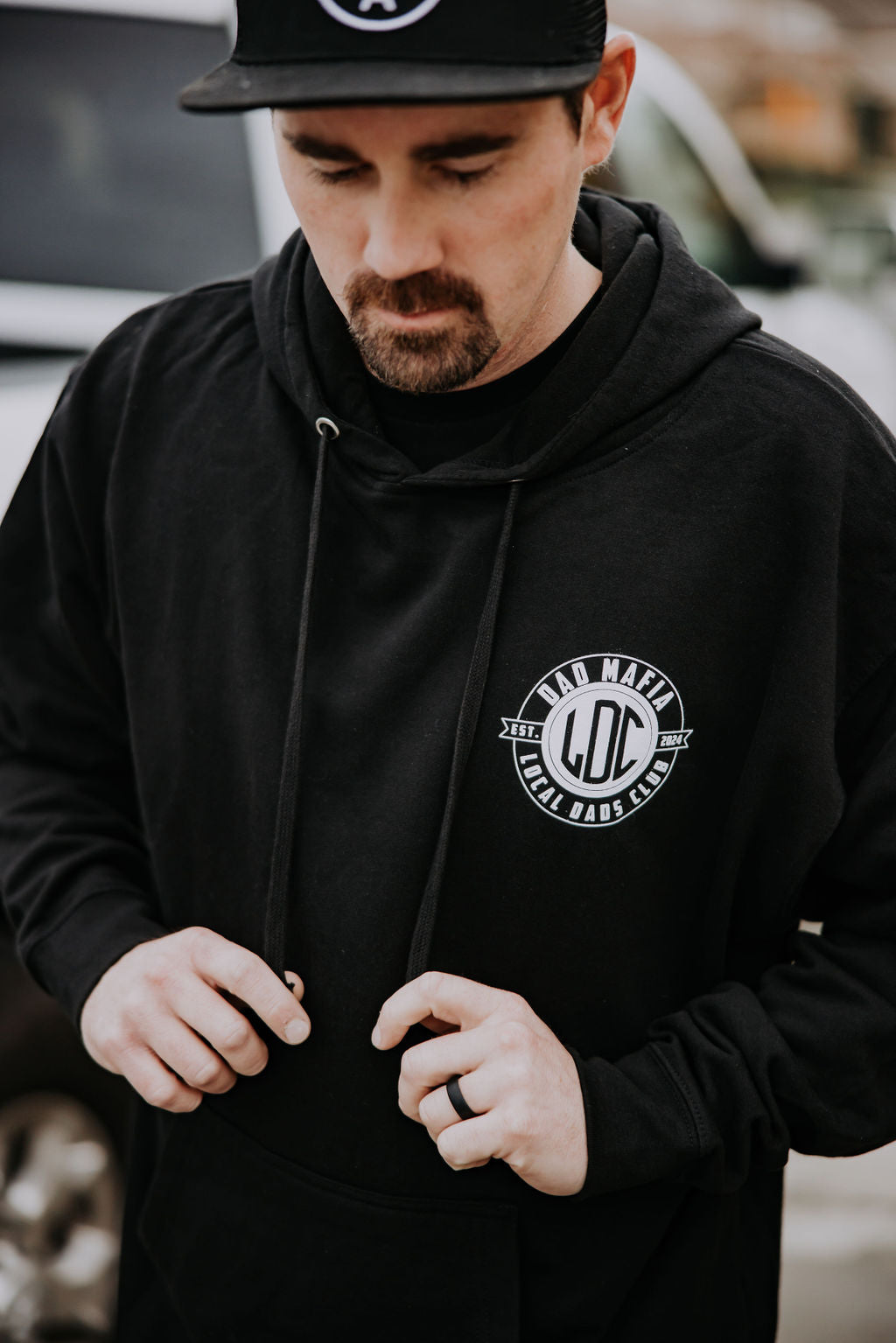 A man wearing a black hoodie and matching cap looks down while adjusting the hoodie’s drawstrings. The hoodie has a "Dad Mafia Lil Dads Club" logo on the chest.
