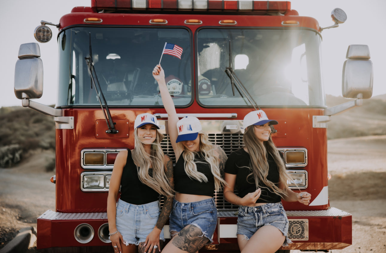 Three women in matching hats and black tops pose in front of a red fire truck, with the woman in the center holding a small American flag and smiling.