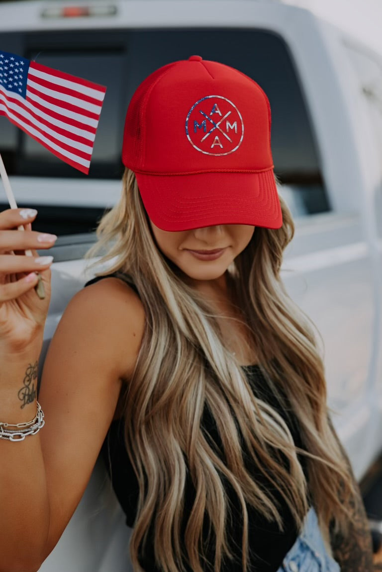A woman with long blonde hair wearing a red "MAMA" hat and black tank top stands by a white truck, holding a small American flag. Her face is partly hidden by the hat's brim.