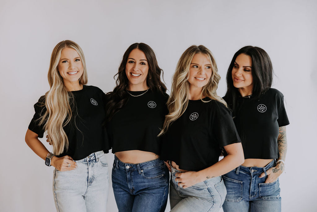 Four women stand side by side, smiling and wearing matching black crop tops and blue jeans, against a plain white background.
