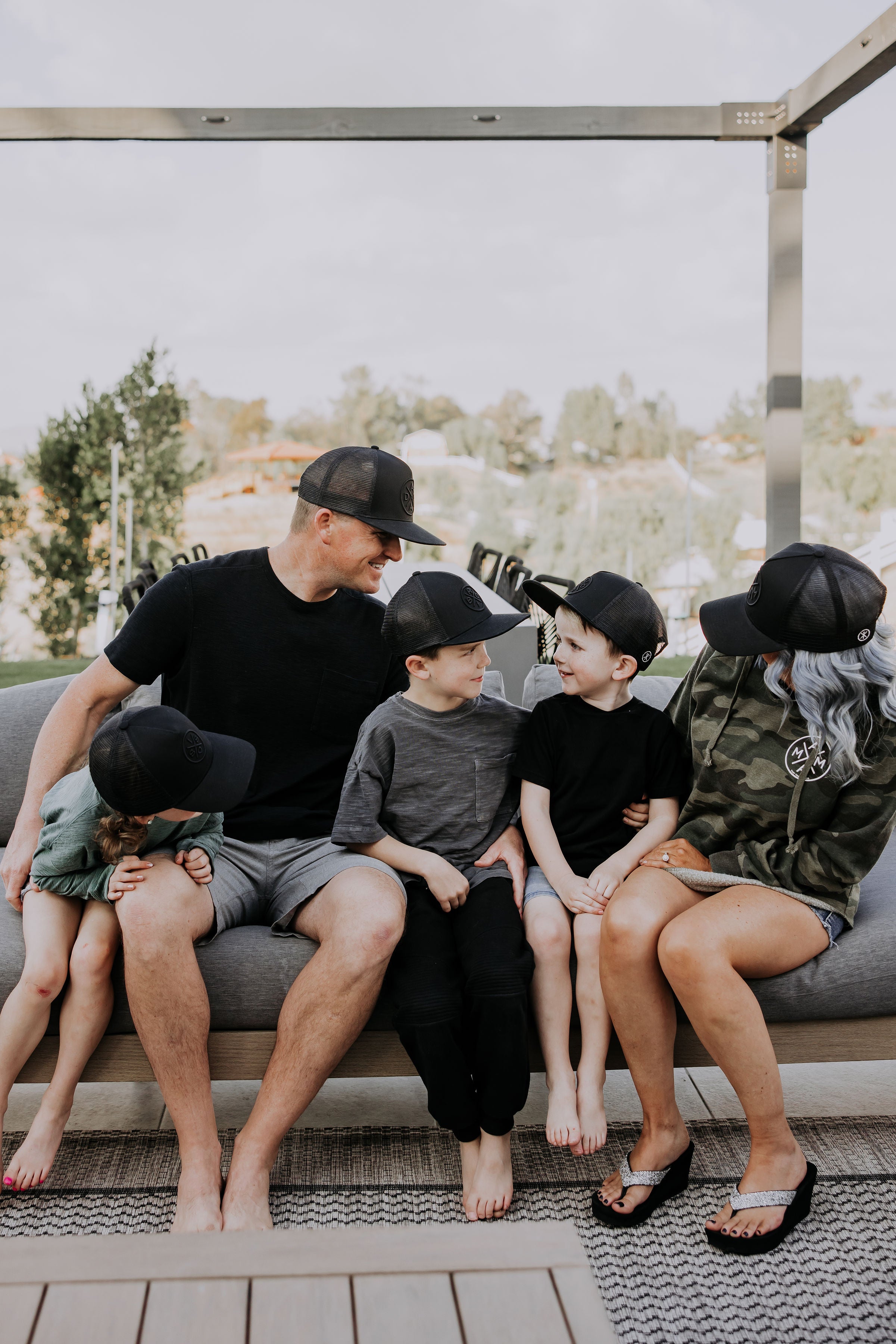 A family sits on an outdoor couch wearing matching black baseball caps. The dad and mom sit at each end, with their three young children between them, smiling and interacting with each other. Trees and houses are visible in the background.