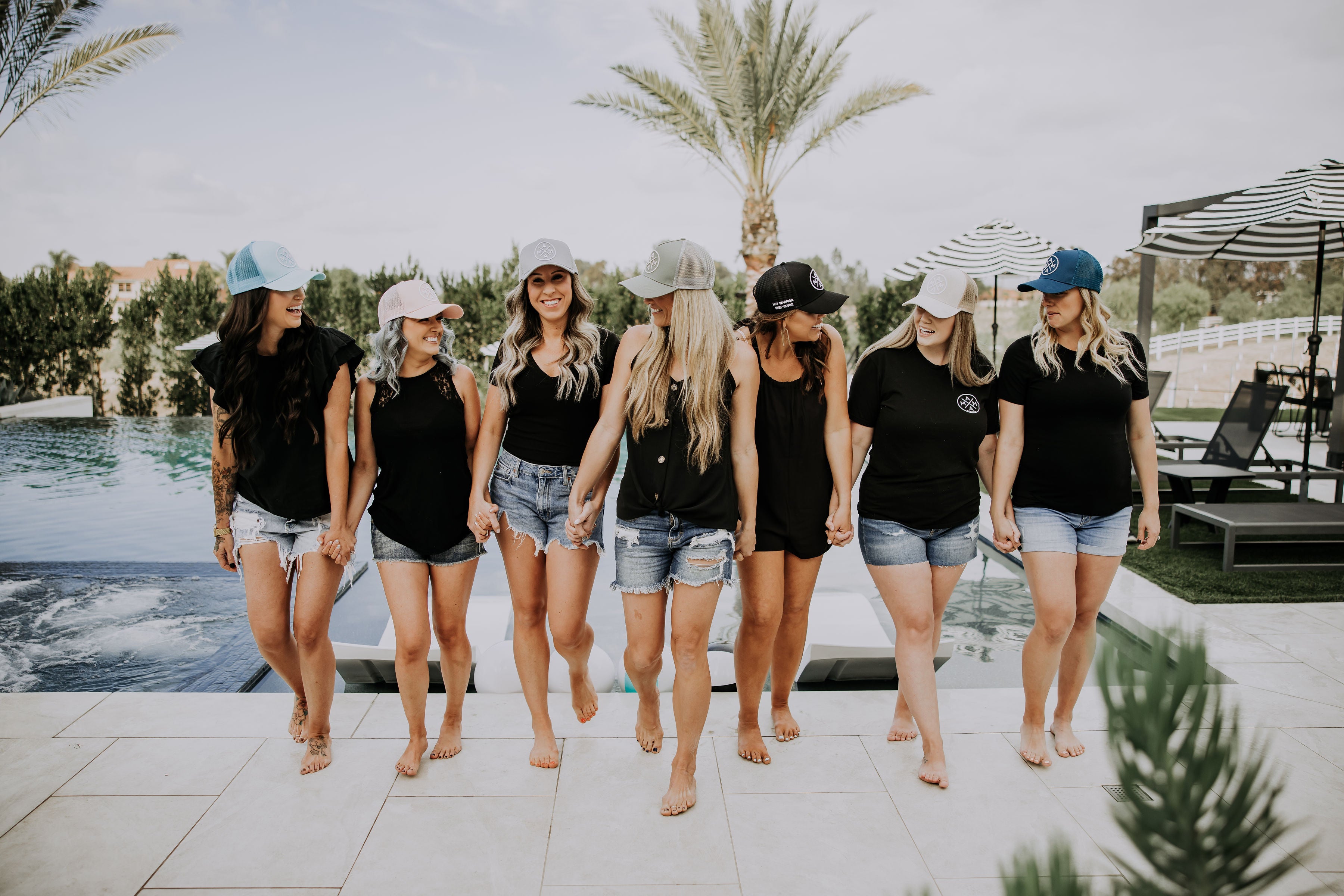 A group of seven women in casual summer attire walk barefoot by a pool, holding hands and smiling. They are wearing shorts, black tops, and various colored baseball caps. Palm trees and lounge chairs are in the background.