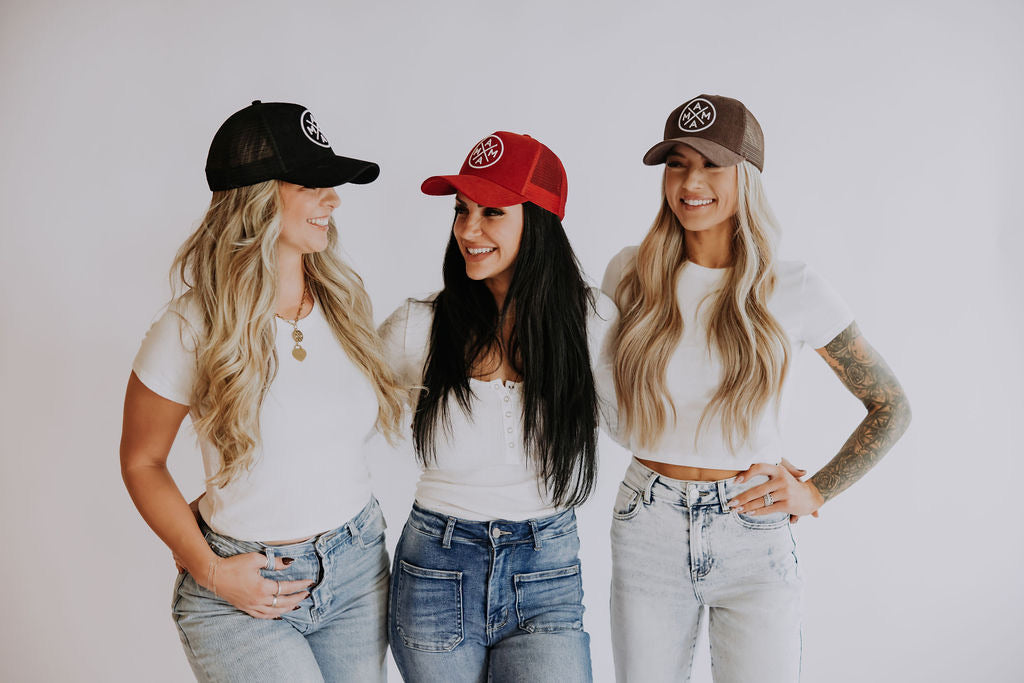 A smiling woman with long dark hair wearing a black T-shirt and black cap holds the cap’s brim with one hand. She has bracelets on her wrist and is posing against a plain white background.