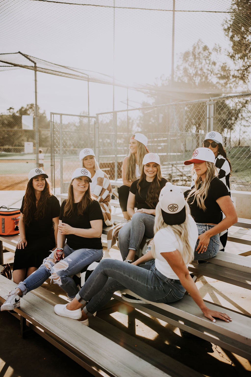 Three women in jeans, white tops, and baseball caps stand together smiling against a plain white background. The woman in the center wears a red cap; the others wear black and brown caps.