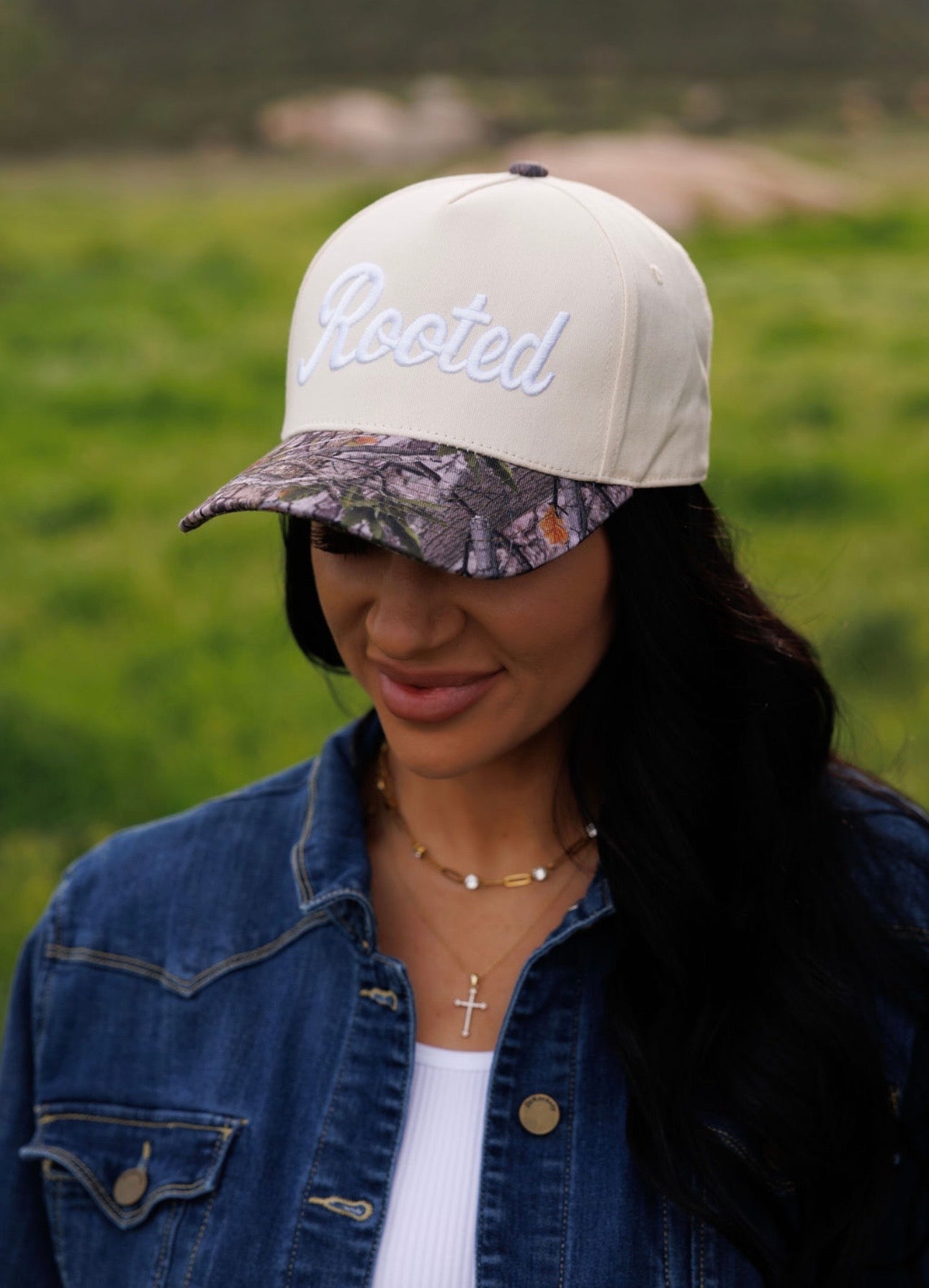 A woman with long dark hair stands in a green field wearing the Mama X™ Brand "Rooted Camo" hat, a denim jacket, white shirt, and cross necklace.