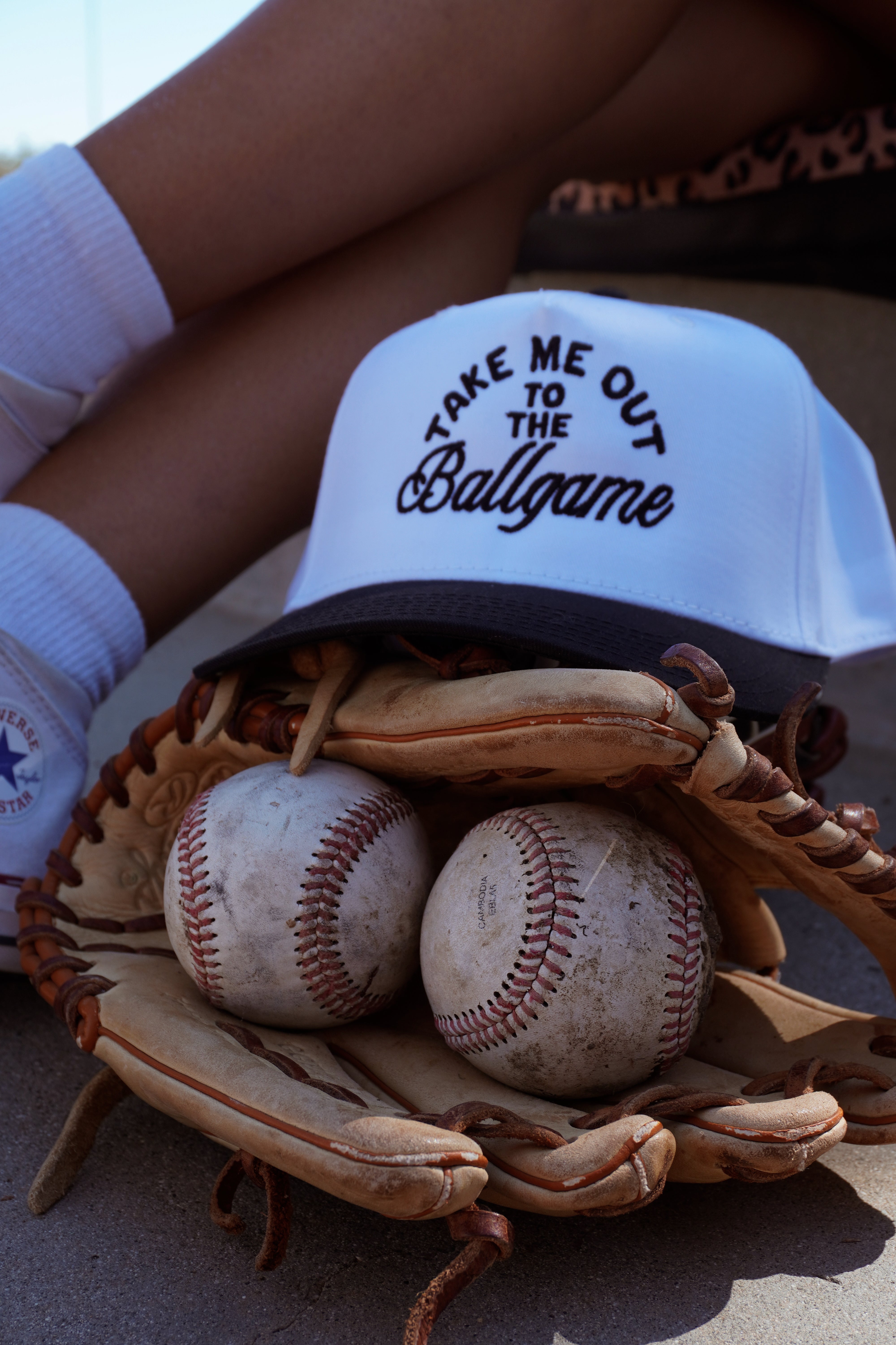 The Ballgame Trucker by Mama X™ Brand—a white and navy hat with "Take Me Out to the Ballgame"—rests on a glove with two worn baseballs, while socked legs and shoes appear in the background.