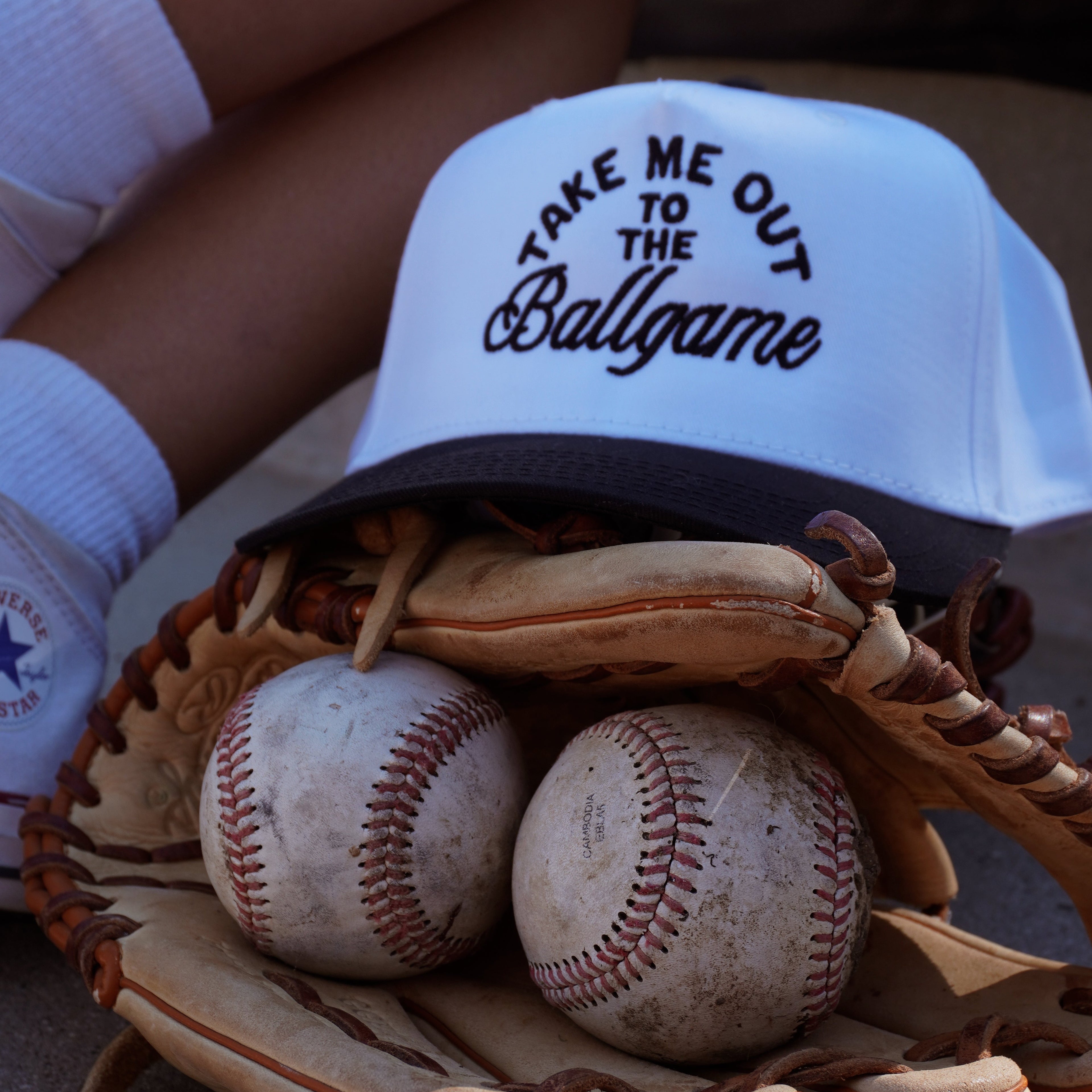 The Ballgame Trucker by Mama X™ Brand—a white and navy hat with "Take Me Out to the Ballgame"—rests on a glove with two worn baseballs, while socked legs and shoes appear in the background.