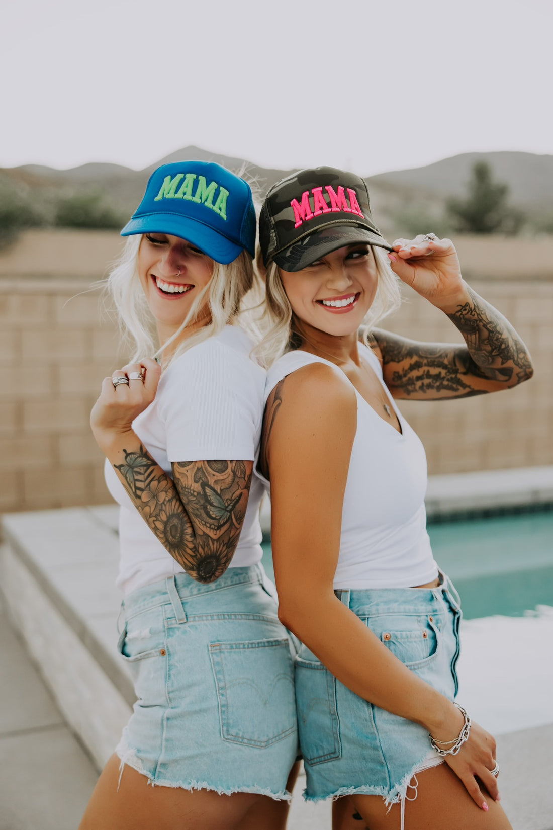Two women with tattoos wear matching Mama X™ Brand Neon Blue/Green Foam Hats, white tops, and denim shorts as they stand back-to-back smiling by a pool with a brick fence and mountains behind them.