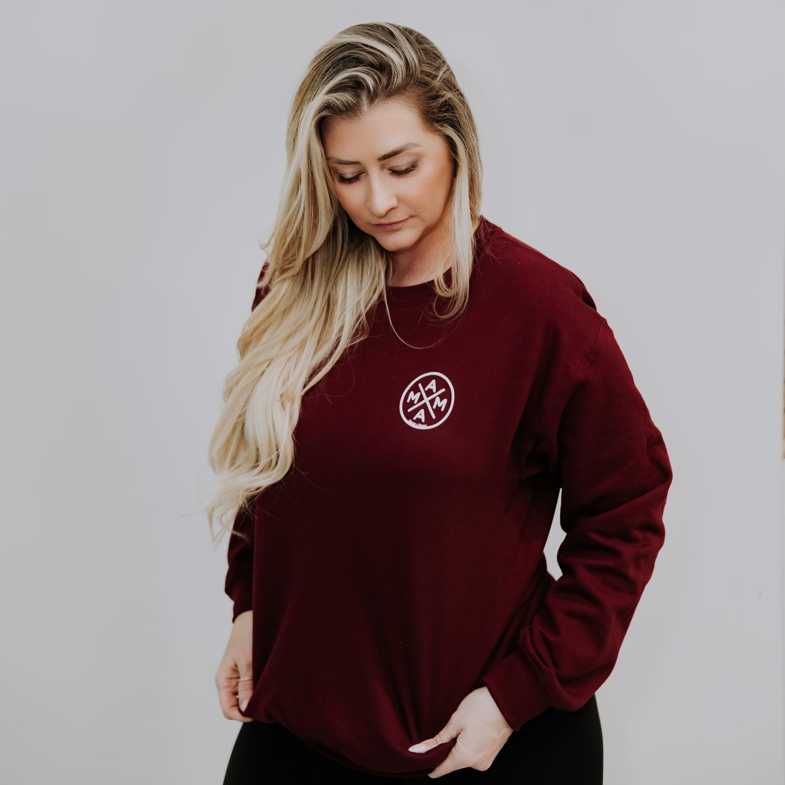 A person with long blonde hair stands against a plain backdrop, looking down with hands in pockets, wearing the Mama X™ Maroon Crewneck featuring a small, round Mama X logo on the chest.