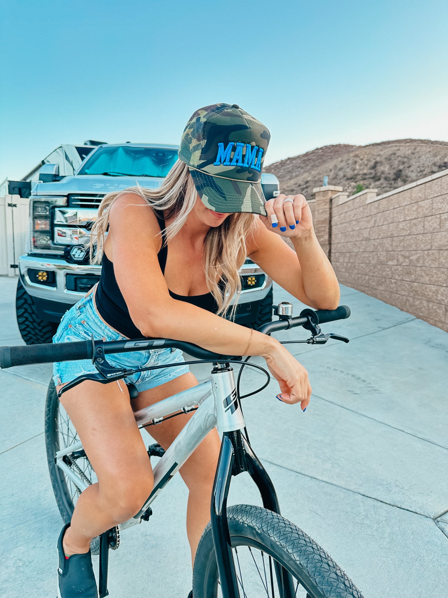 A woman in a black tank top sits on a bicycle, holding the brim of her Mama X™ Brand Camo/Blue Mama Foam Hat. A large truck, desert hills, and a stone wall are visible in the background.