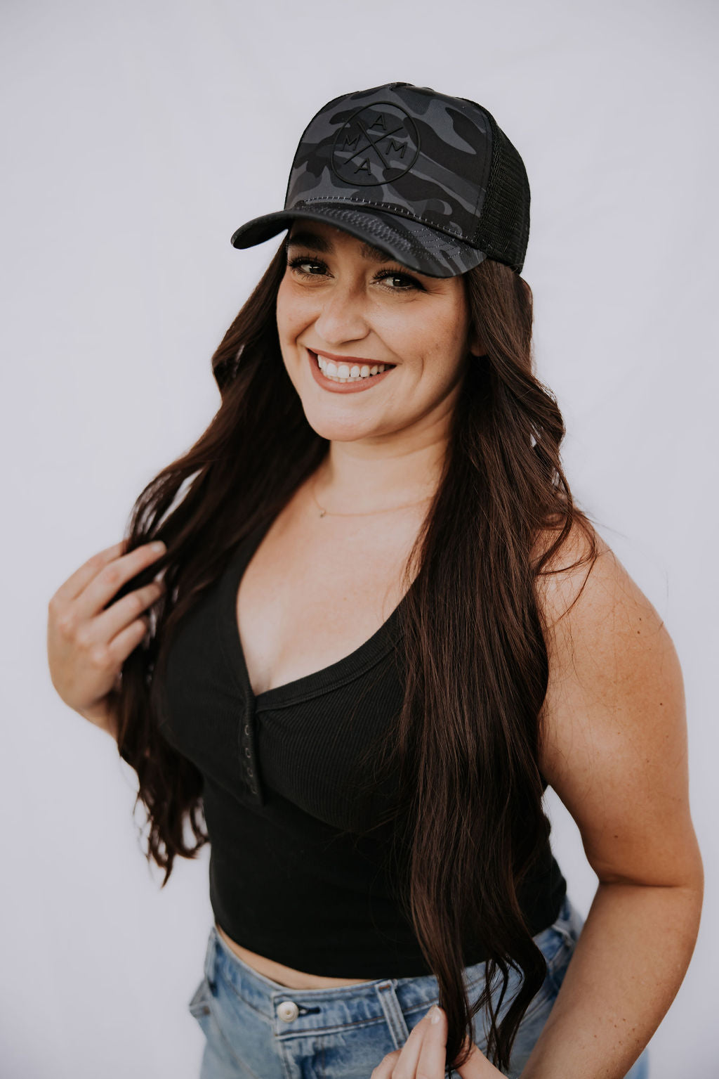 A smiling person with long dark hair is photographed against a plain white background wearing a black sleeveless top, denim jeans, and the stylish Black Camo Mama X™ Trucker hat by Mama X™ Brand, featuring an adjustable snapback closure.