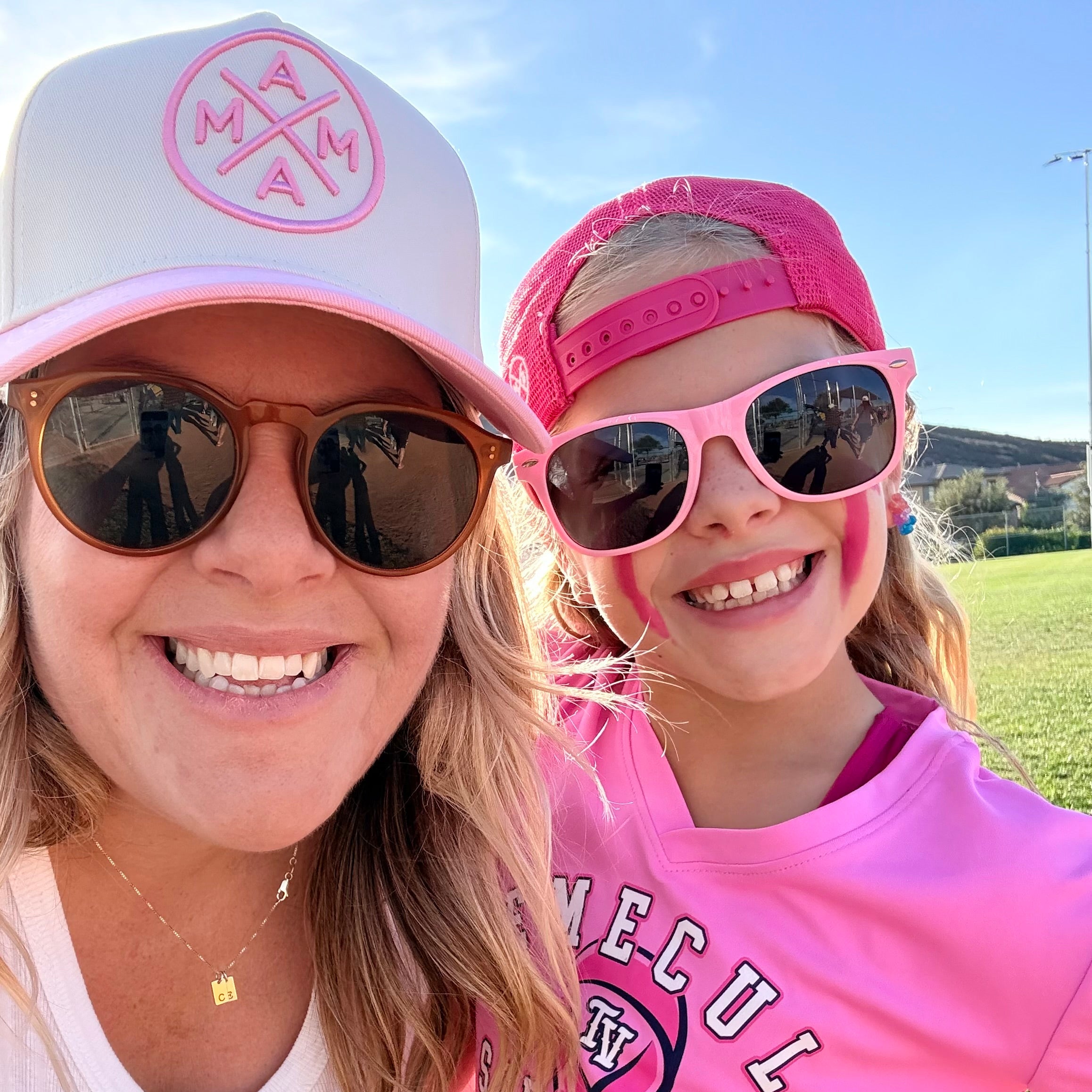 A woman and a young girl in sunglasses smile outdoors on a sunny day. The girl wears a Temecula Softball shirt and the Pink Mama X™ Baseball Cap by Mama X™ Brand, with a grassy field and blue sky making for a cheerful scene.