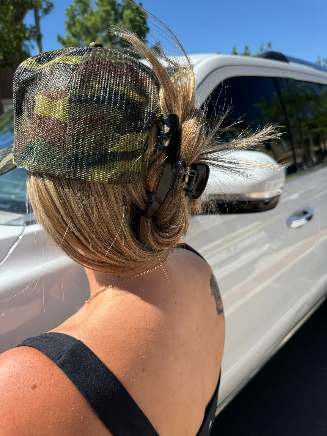 A person with blonde hair in a messy bun, secured by the Mama X™ Brand Black Trucker Claw Clip, wears a camouflage cap and black tank top while standing next to a white SUV on a sunny day.