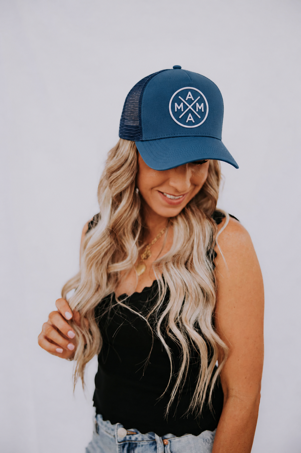 A woman with long, wavy blonde hair wears a black camisole, light denim jeans, and the Mama X™ Brand Dark Blue Mama X Trucker hat featuring "MAMA" in a circle design. She smiles and looks down against a plain white background.
