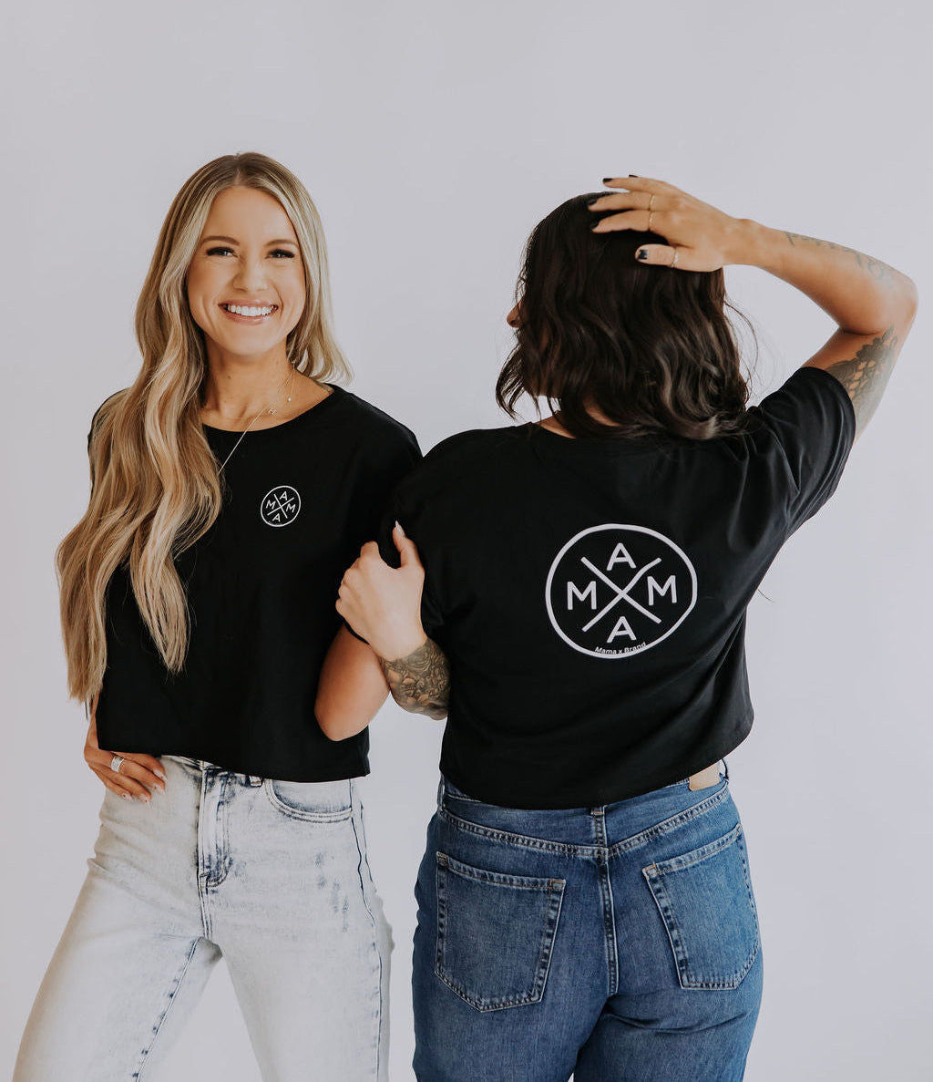 Two women wear relaxed fit Mama X crop tops by Mama X™ Brand with a white circular logo on the back; one smiles at the camera with a hand in her pocket, while the other faces away. Both pair the tops with jeans against a plain background.