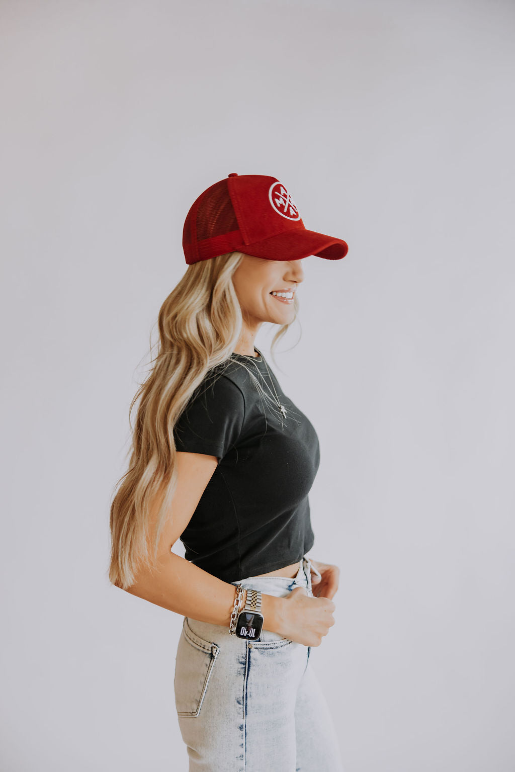 A woman with long, wavy blonde hair smiles in profile against a plain background, wearing the Red Mama X Corduroy Trucker by Mama X™ Brand, paired with a black cropped t-shirt and light-wash jeans.