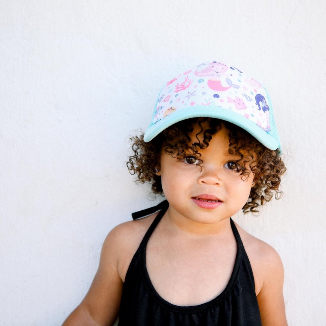 A young child with curly hair stands against a white wall, wearing the Mermaid Trucker Hat by Mama X™ Brand and a black sleeveless outfit. The child has a serious expression and gazes directly at the camera.