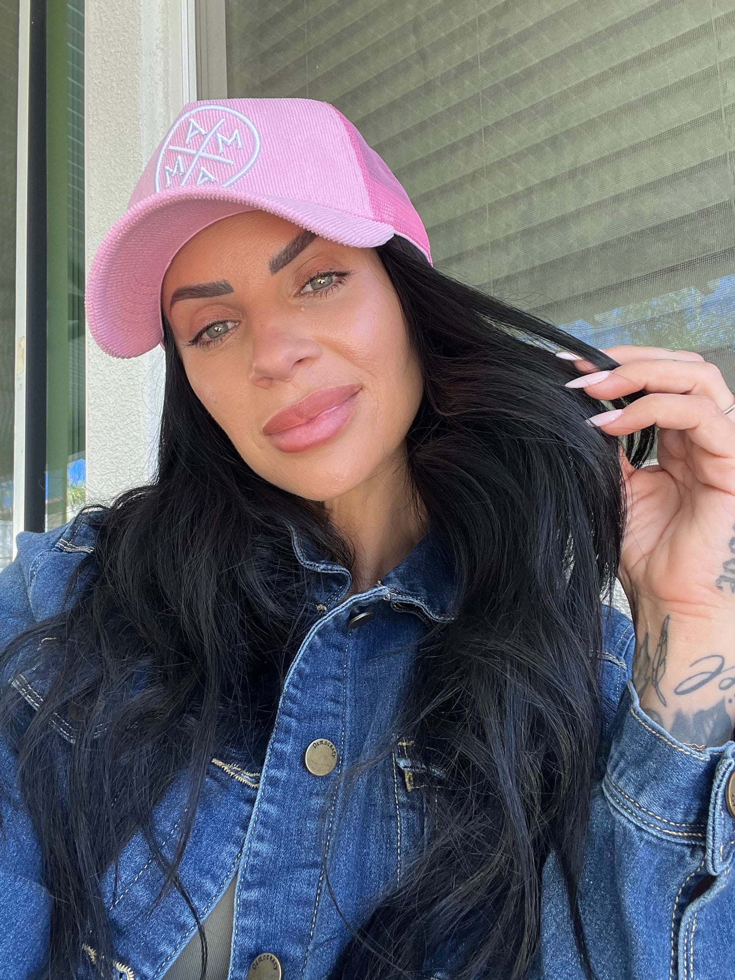 A woman with long dark hair wears the Pink Corduroy Mama X Trucker hat by Mama X™ Brand and a denim jacket, sitting indoors by a window with blinds. She holds a strand of her hair and looks at the camera with a slight smile.
