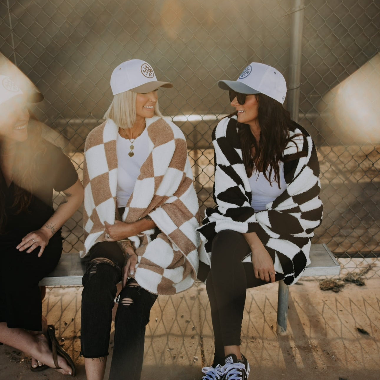 Two friends sit on a bench, wrapped in butter-soft Mama X Checkered Blankets and wearing white caps. One sports a beige blanket with white shoes, the other a black blanket with black shoes. They smile and chat under sunlight filtering through the fence—a cozy gift of friendship from Mama X™ Brand.