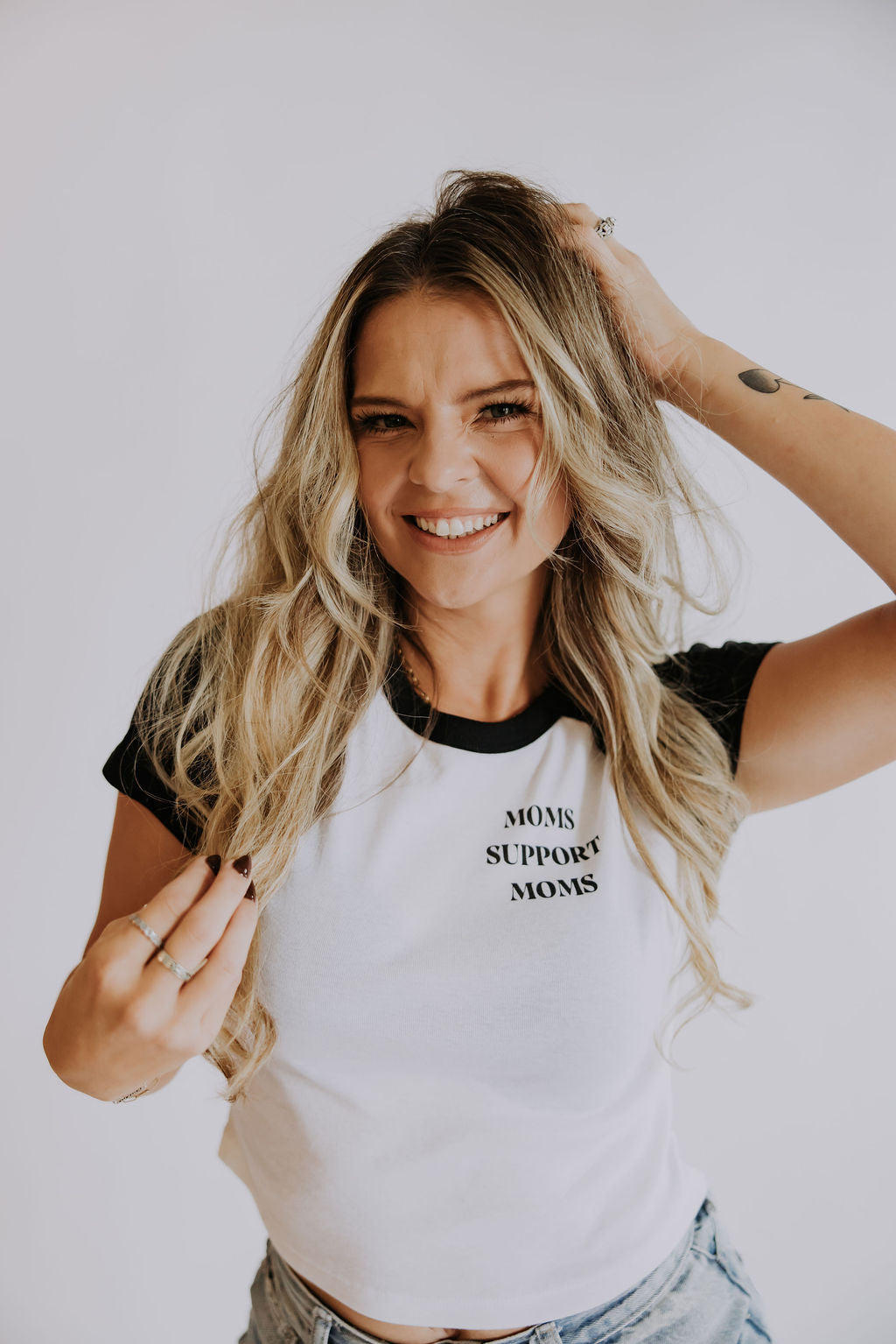 A smiling woman with long blonde hair wears the Mama X™ Brand "Moms Support Moms" raglan baby tee and poses against a light background, holding her hair and smiling at the camera.