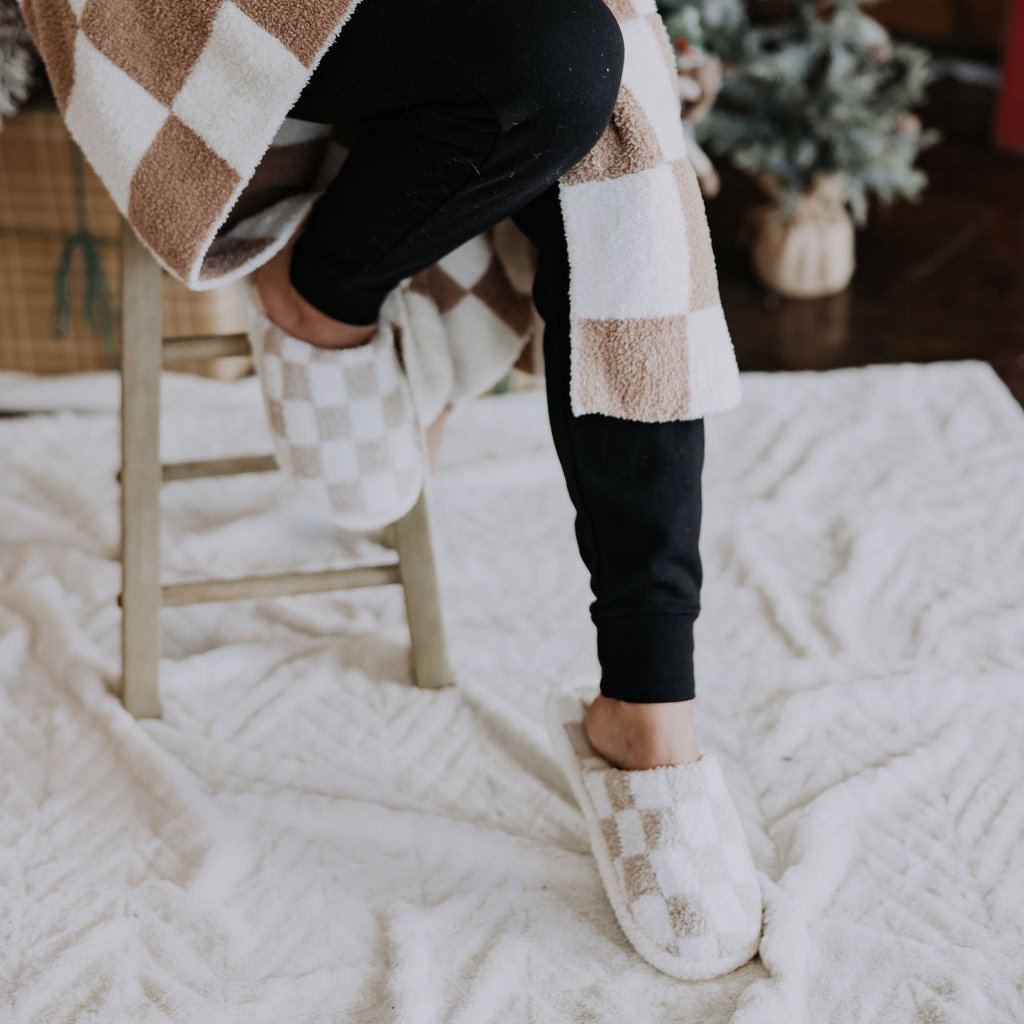 A person sits comfortably on a stool, enveloped in a checkered blanket and wearing Mama X Check Board Slippers. Their legs are covered by black pants. In the background, a small, decorated Christmas tree stands on a white textured rug, with the subtle Mama X™ Brand logo adding an elegant touch to the cozy scene.