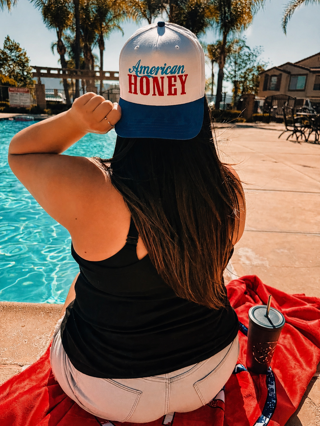A woman sits by a pool on a red towel, wearing the Mama X™ Brand "American Honey" cap, black tank top, and light shorts. With her long hair down, she holds the cap&