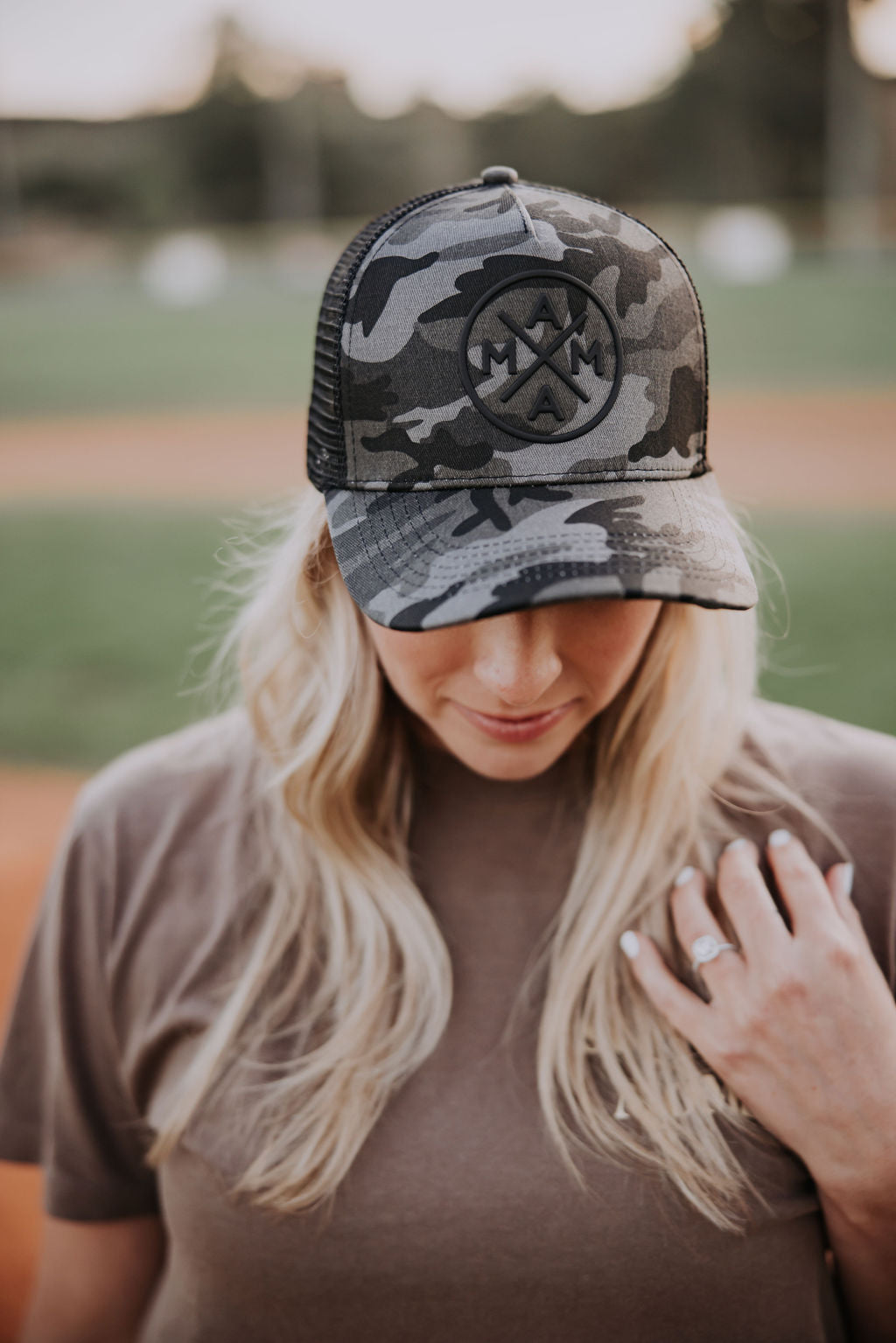 A person with long blonde hair wears a Black Camo Mama X™ Trucker from Mama X™ Brand, featuring a breathable mesh back and adjustable snapback. They hold the brim down while their other hand rests on their shoulder, standing outdoors on a dirt field.