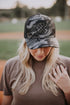 A person with long blonde hair wears a Black Camo Mama X™ Trucker from Mama X™ Brand, featuring a breathable mesh back and adjustable snapback. They hold the brim down while their other hand rests on their shoulder, standing outdoors on a dirt field.