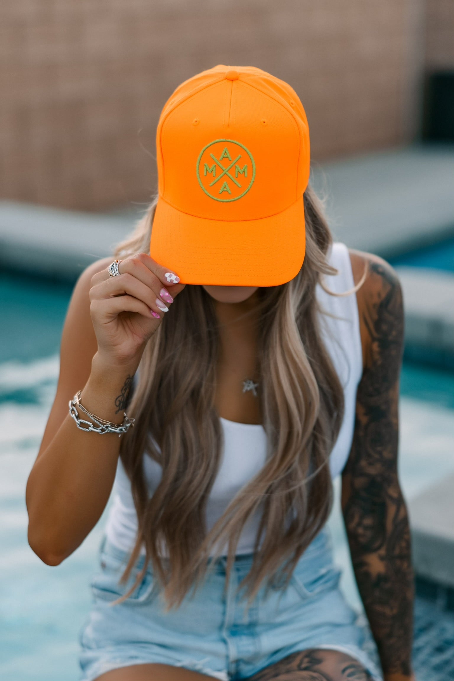A woman with long wavy hair and tattoos, in a white tank top, denim shorts, and the Neon Orange Mama X hat by Mama X™ Brand, sits poolside holding the brim to partially cover her face.